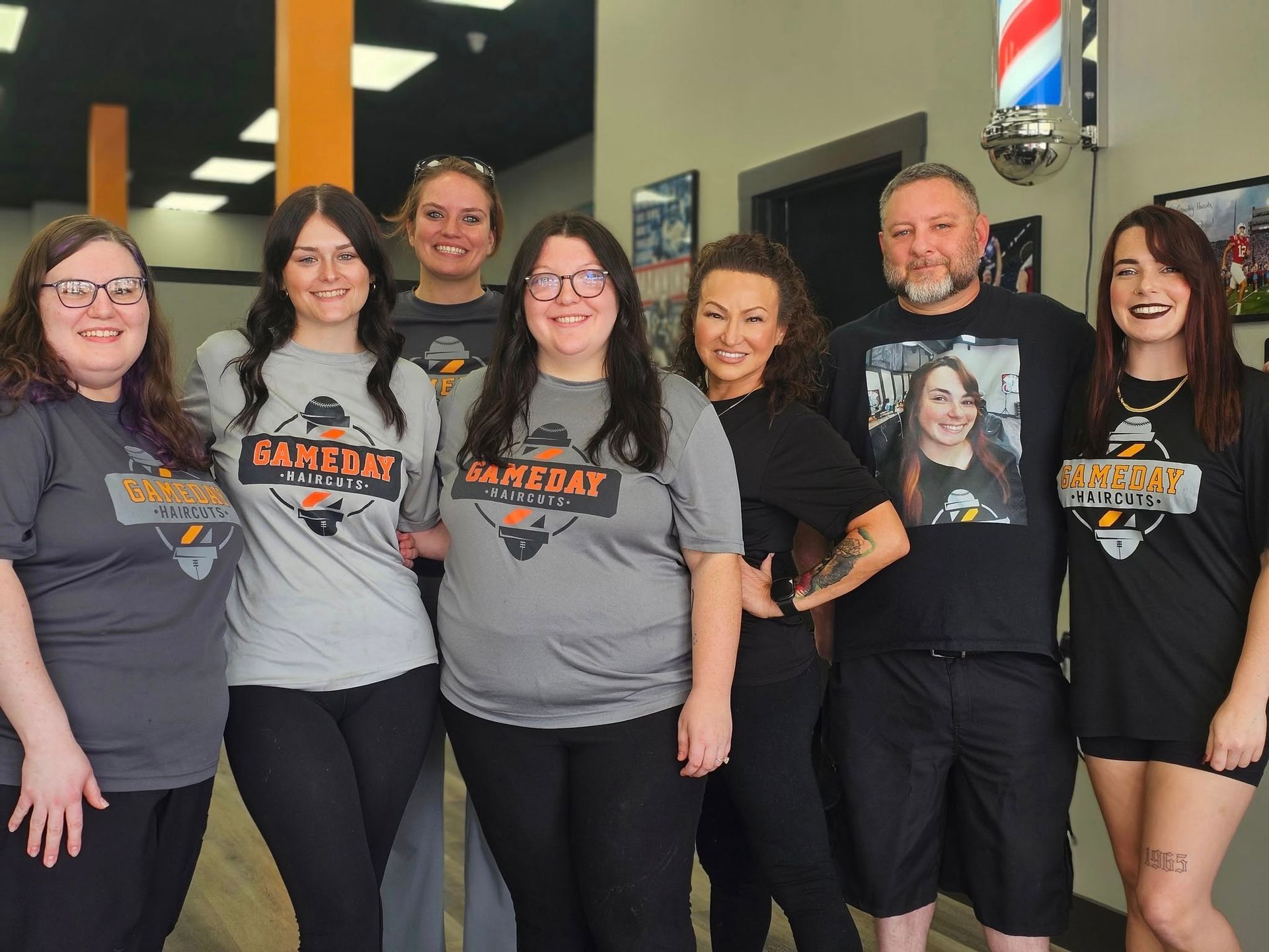 Group of people in a barbershop wearing matching "GameDay Barber Co." shirts, smiling.