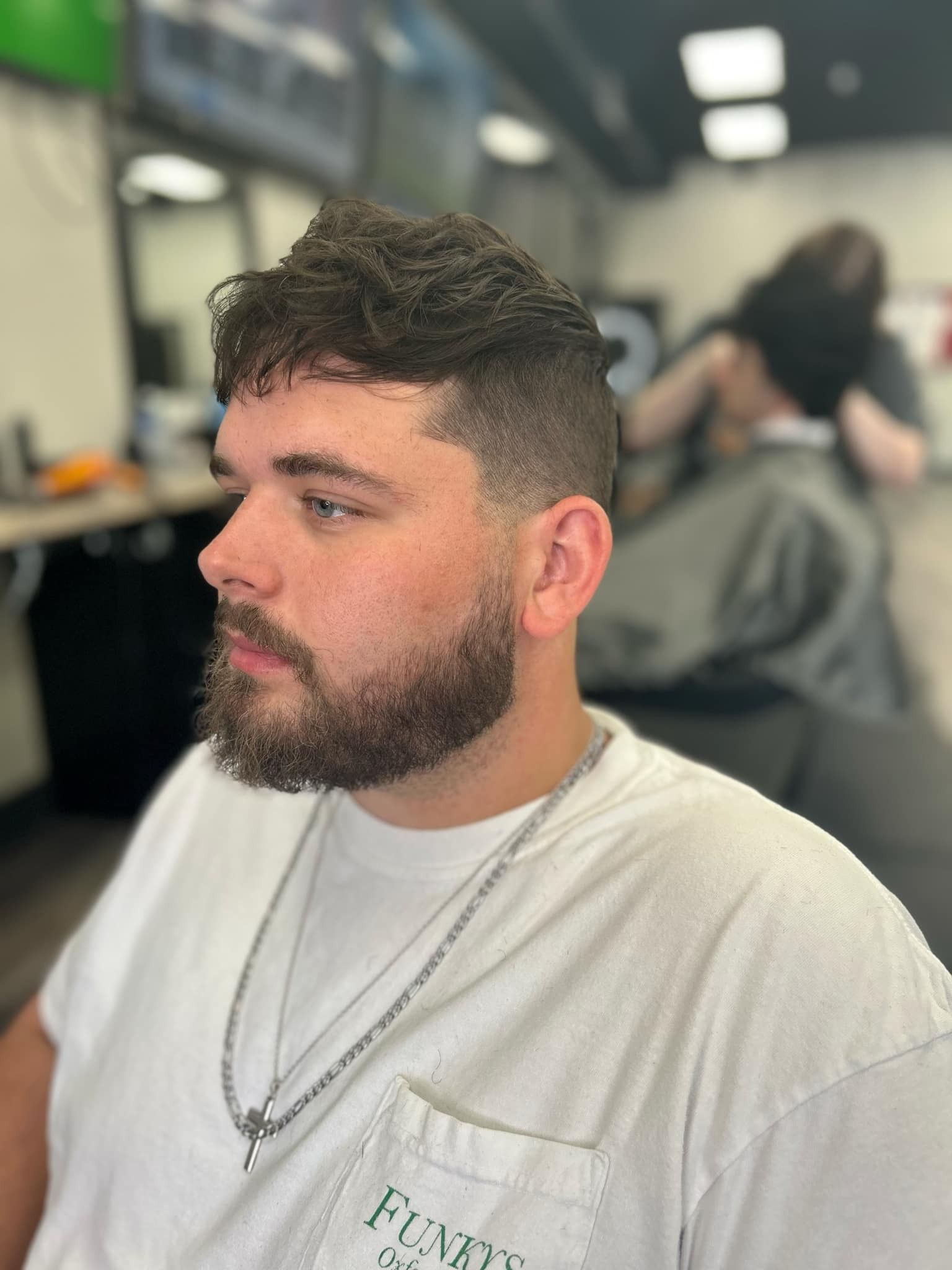 Bearded man in a white T-shirt and silver chains sitting in a blurred barbershop chair.