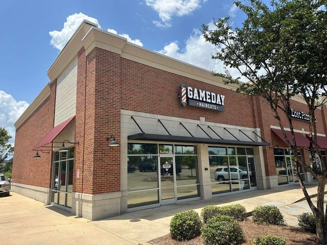 Exterior view of the "Gameday Barber Shop" in a brick building with a dark awning and glass windows on a sunny day.