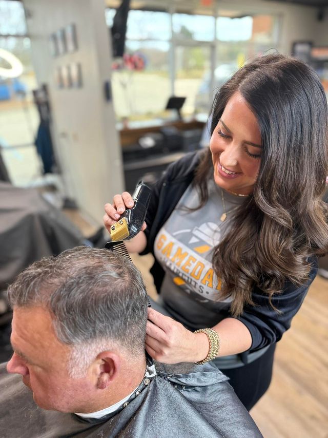 Woman cutting a man's hair in a barber shop. She smiles, holding clippers near his head.