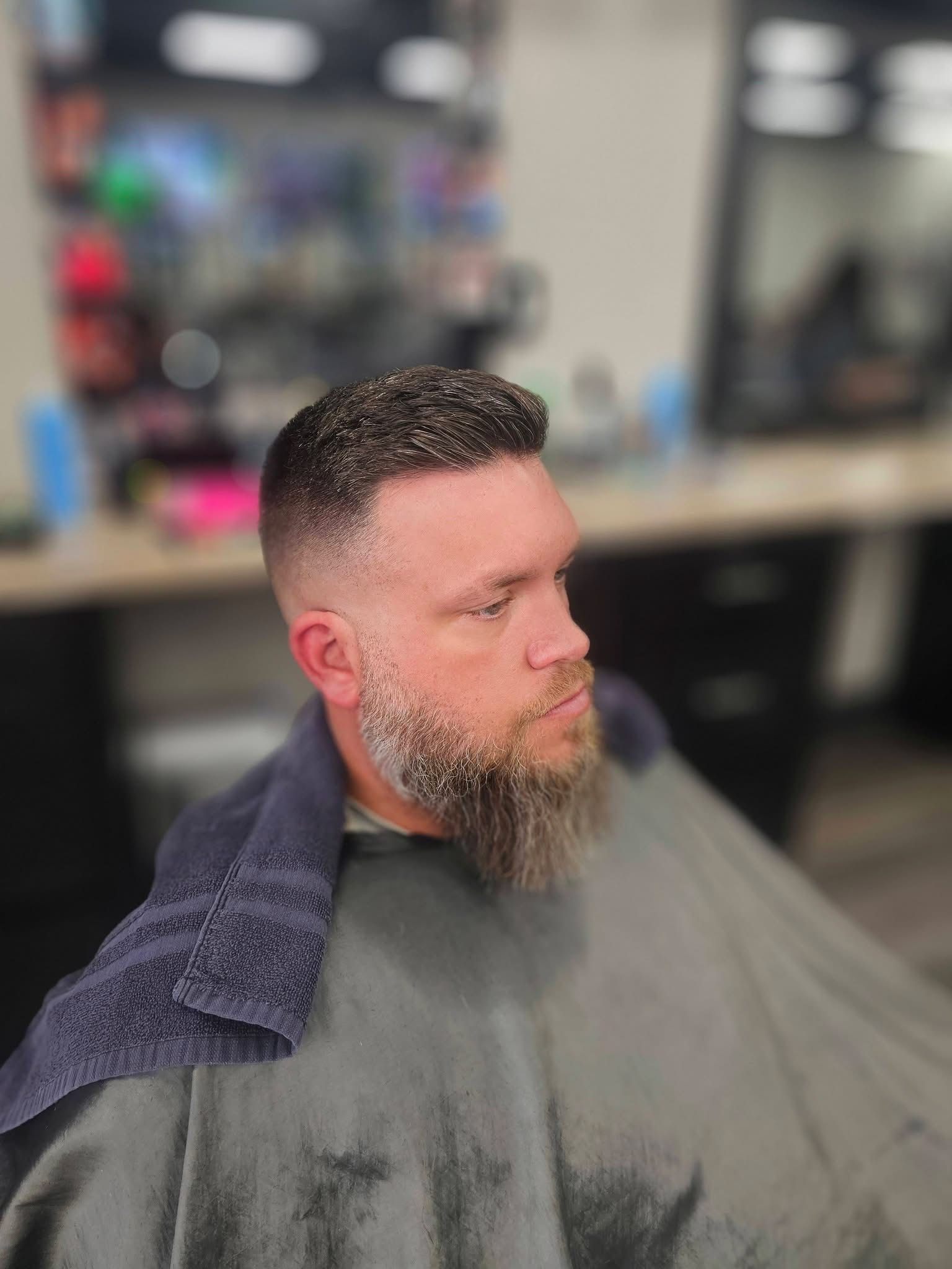Man with short curly hair and beard getting a haircut in a salon, draped in a gray cape