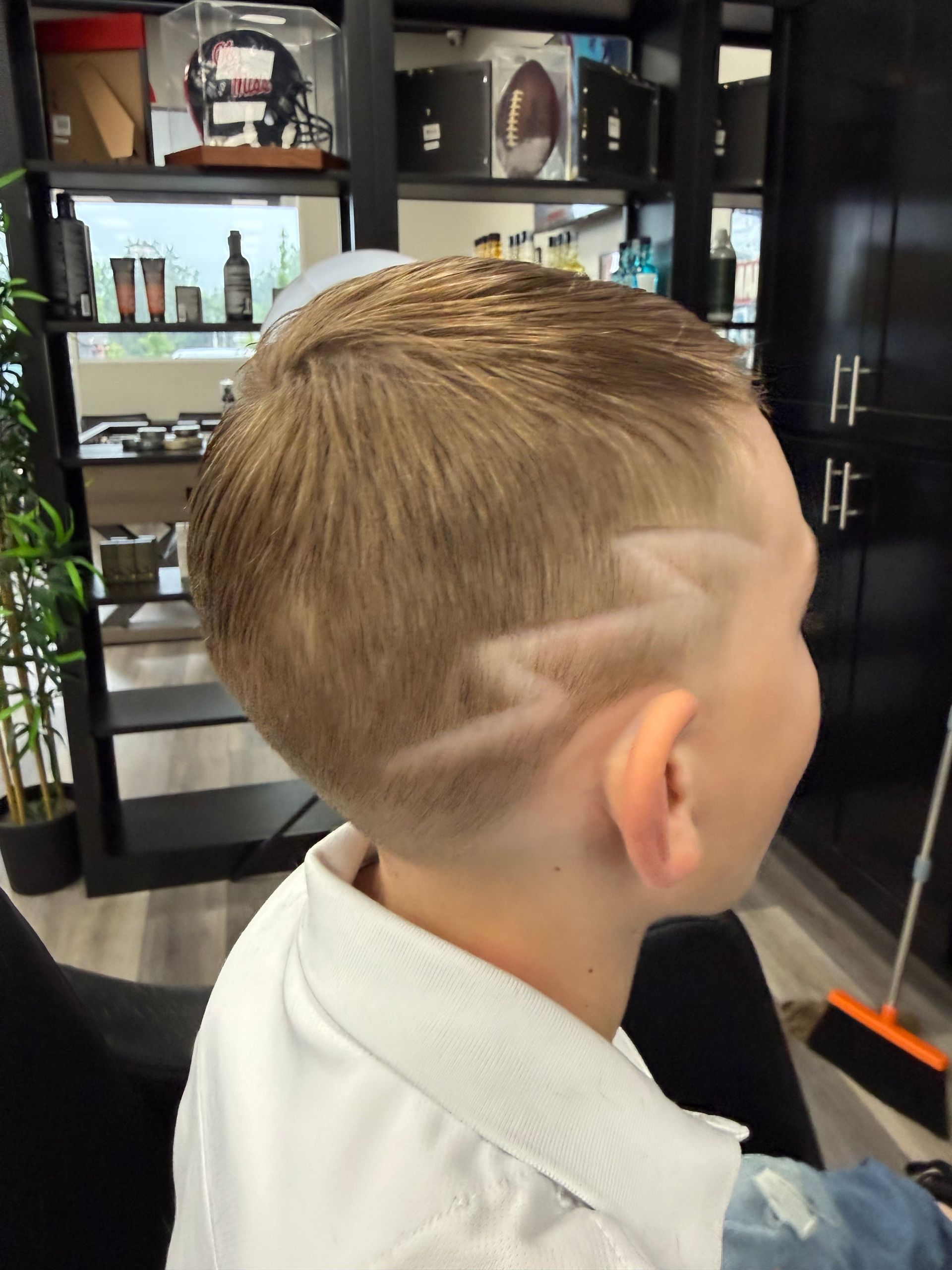 Child with a short blond haircut and shaved design at a barbershop chair, seen from the back.