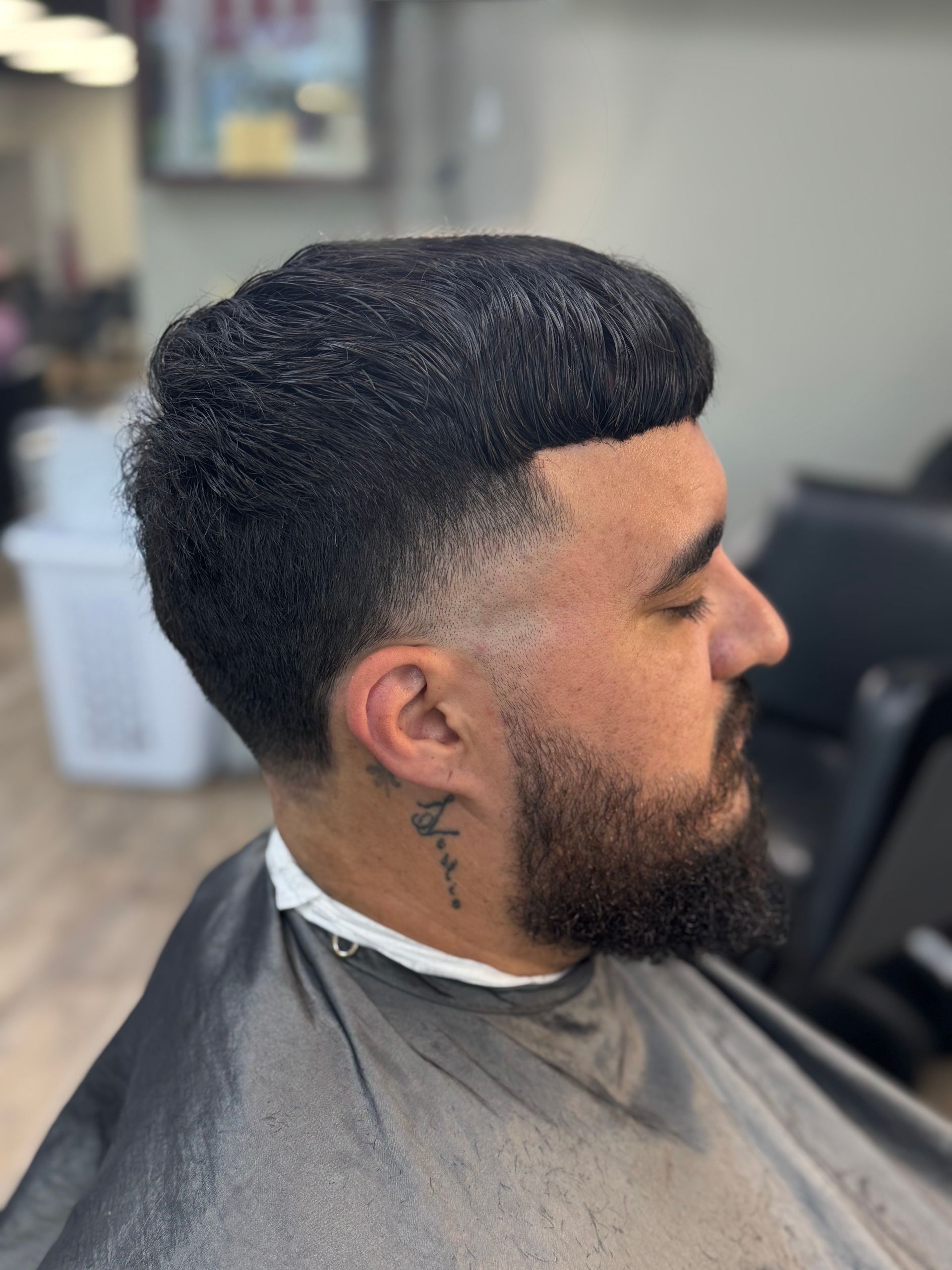 Side profile of a man with a beard and short fade haircut in a barbershop, wearing a haircut cape.