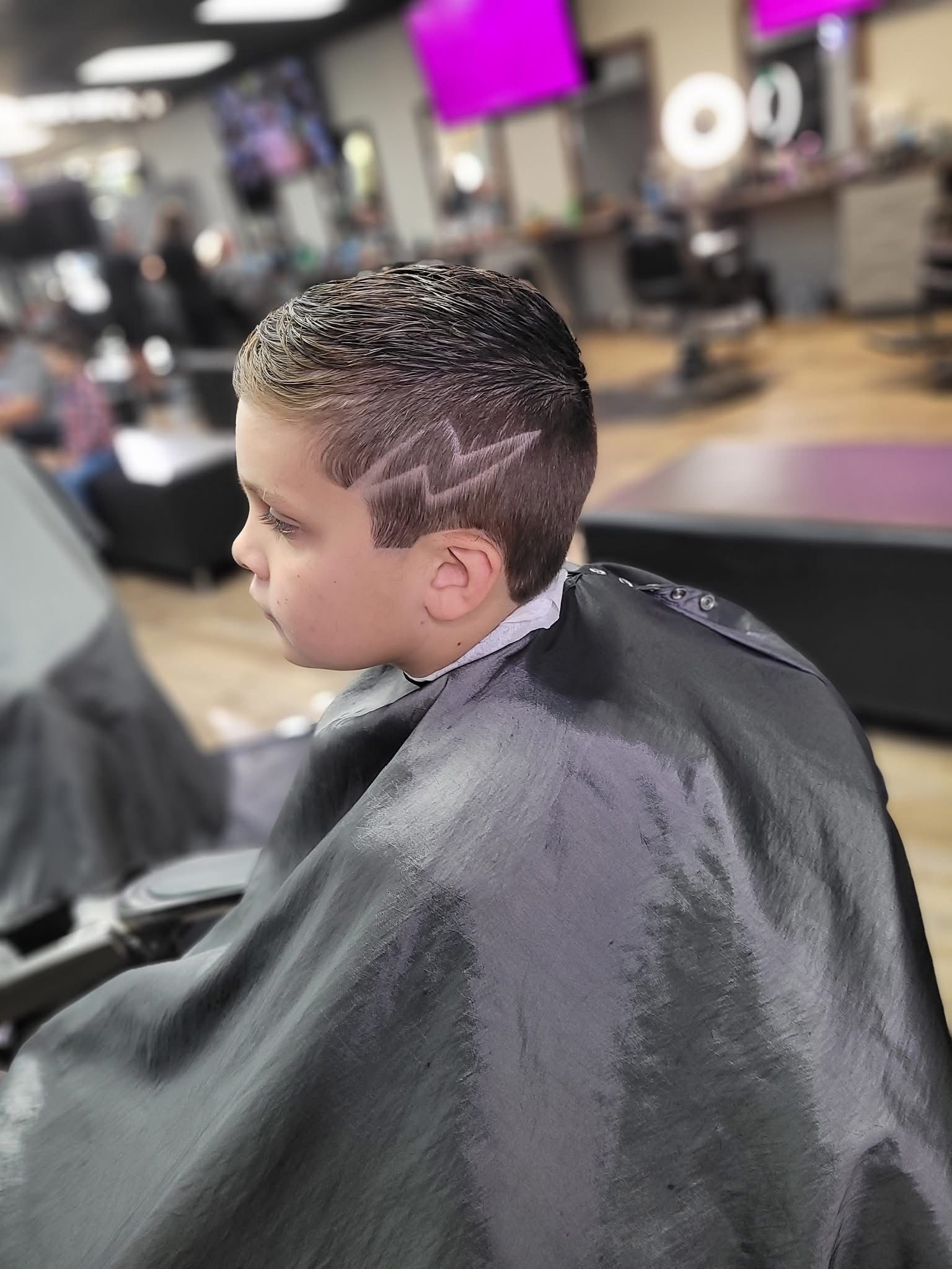 Child with a short haircut and patterned top, seated in a barber chair under a black cape in a salon.