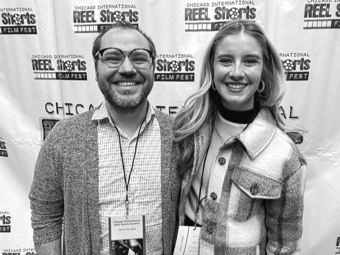 Two people smiling, posing at a film festival. A man with glasses and a woman with long hair stand in front of a banner.