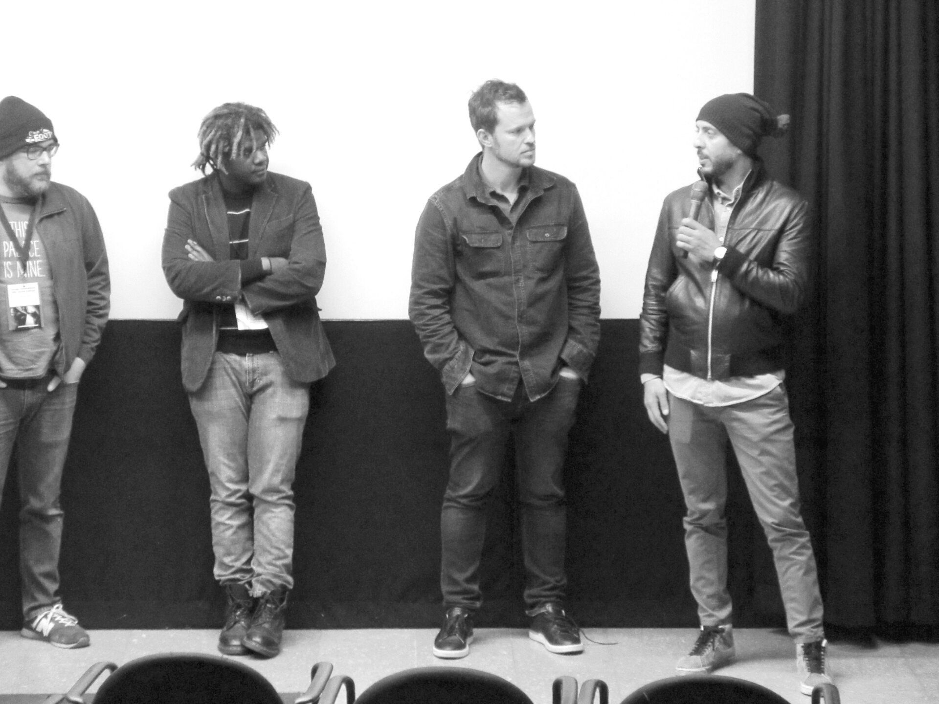 Four men standing on a stage. One holds a microphone. Black and white.