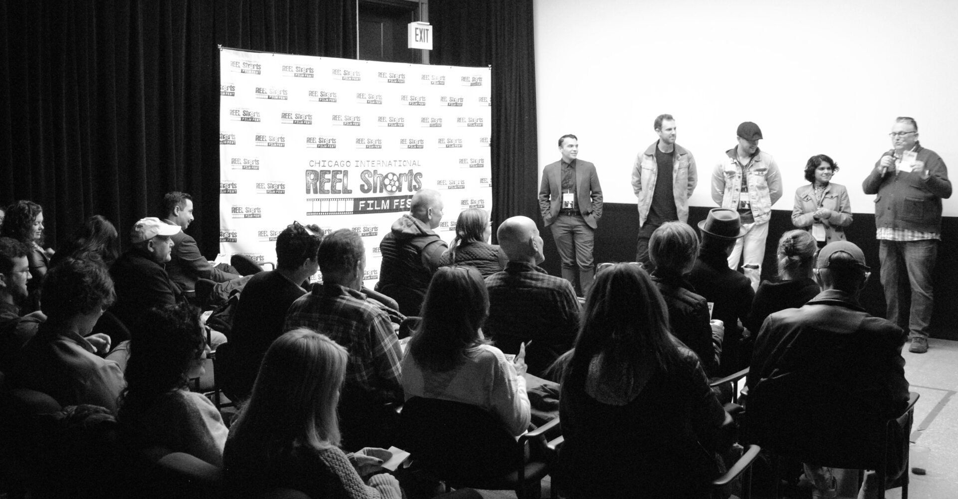 Audience watches panel of speakers on a stage. Backdrop with logo. Black and white photo.