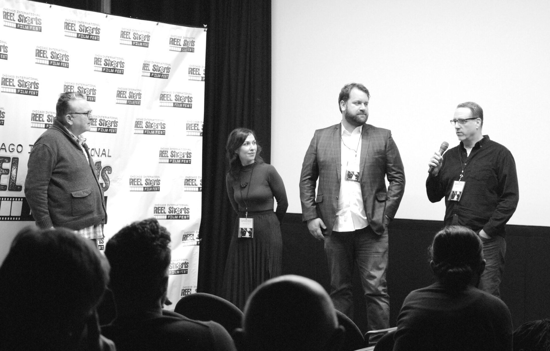 Four people on stage; woman in a dark dress, three men with microphones and audience in the foreground.