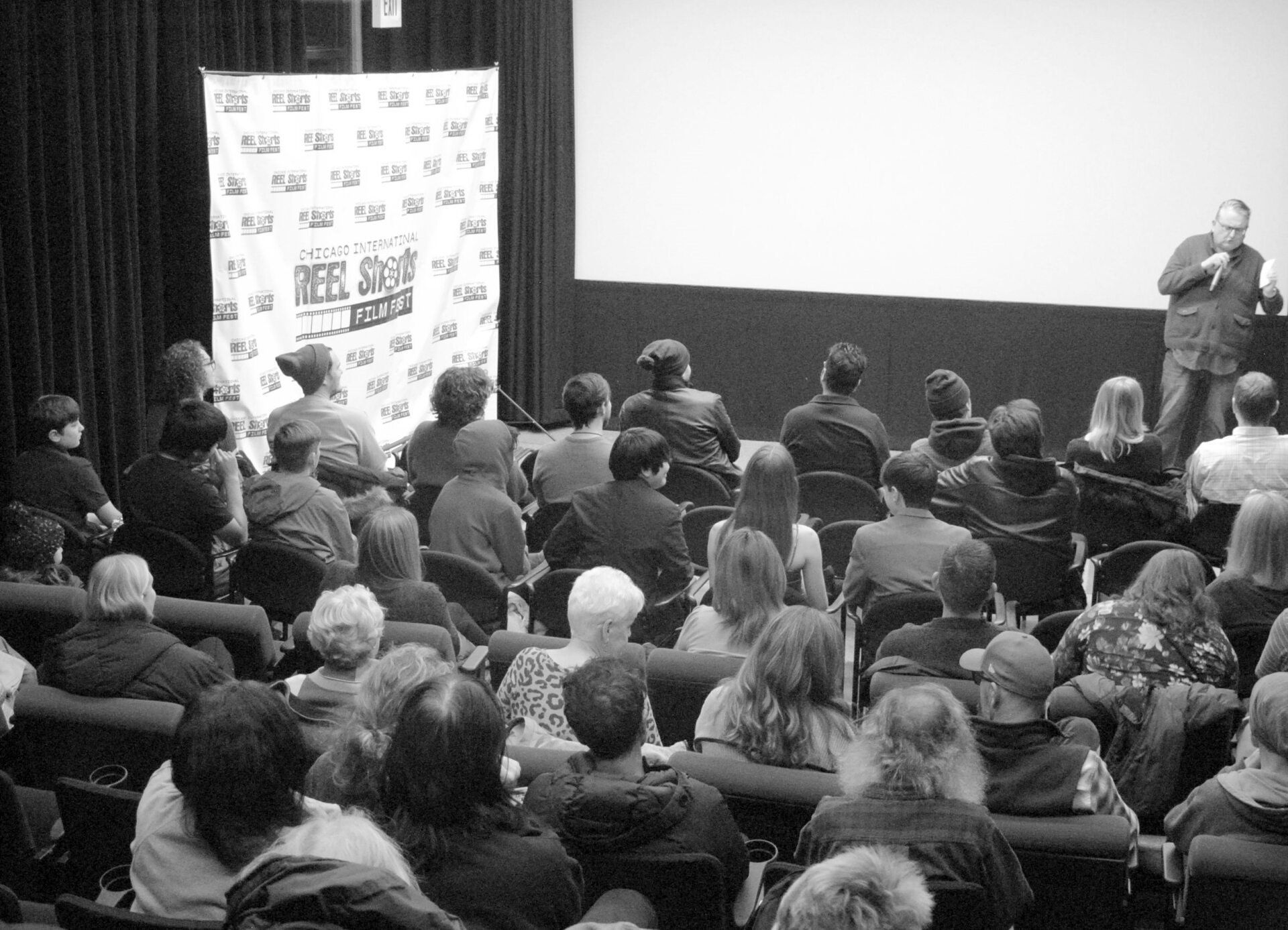 Audience in theater watches a presentation. A man speaks on stage in front of a screen.