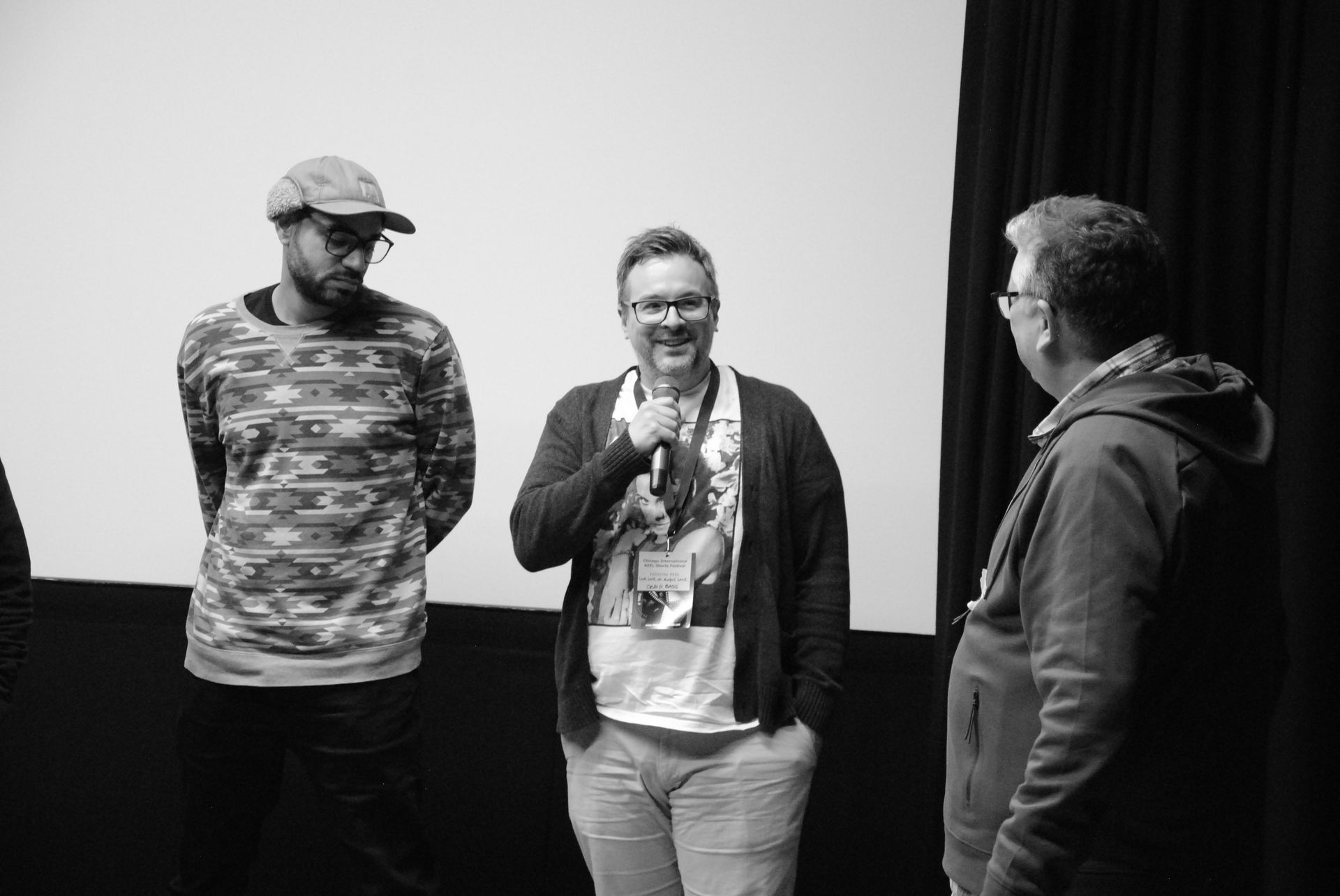 Three men on a stage in front of a screen. One speaks into a microphone. Black and white.