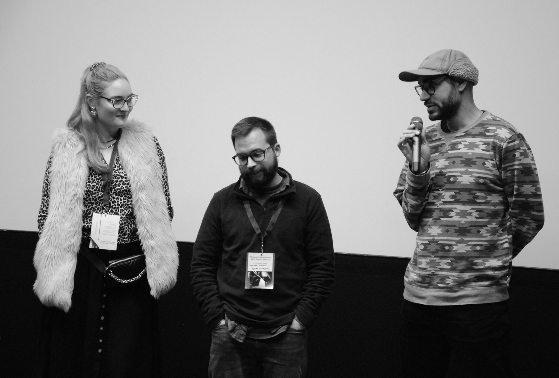 Three people on a stage: a woman in fur vest, a man with a beard, and a man with a microphone.