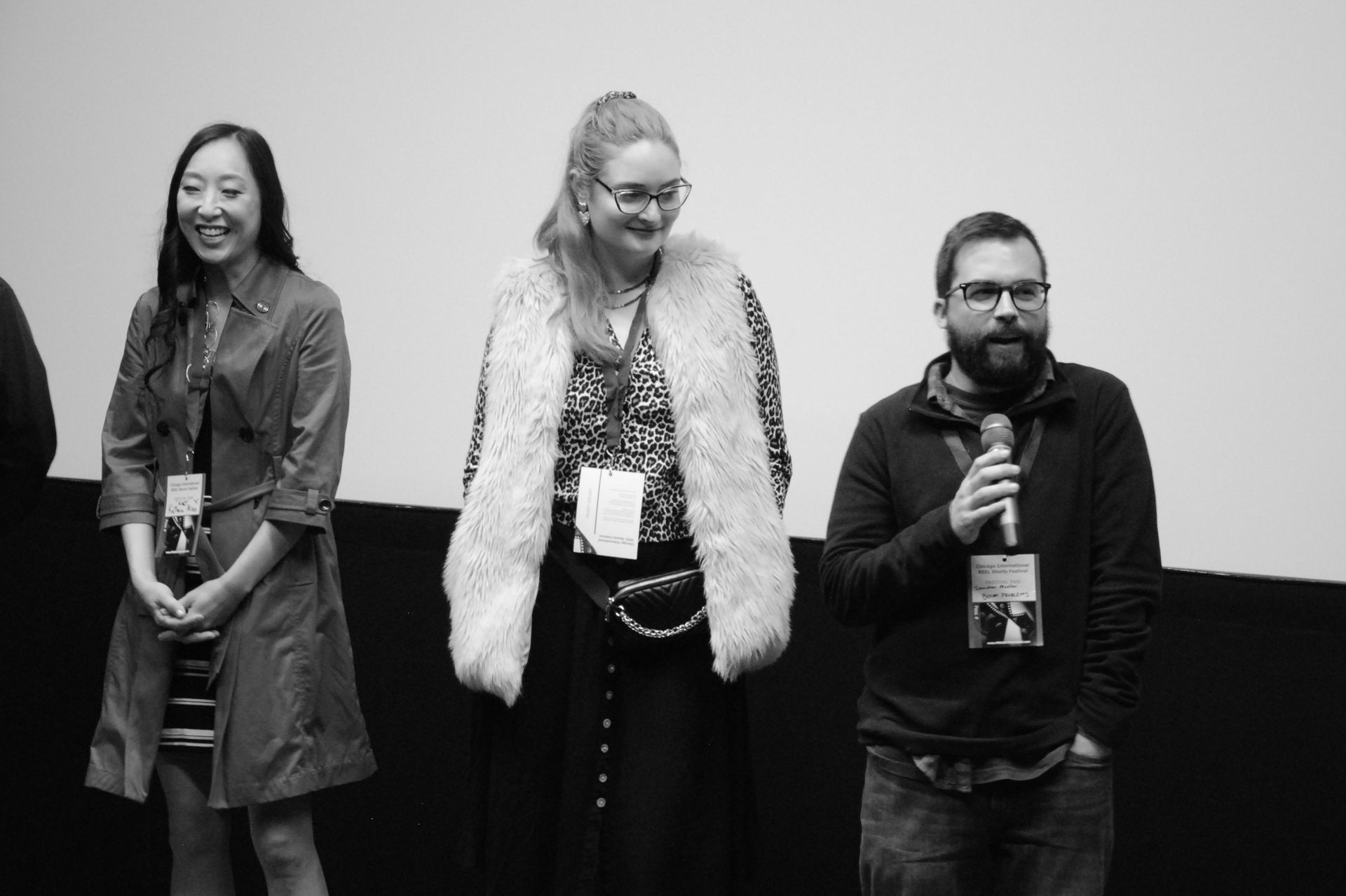 Three people on stage, smiling. Woman in center wears fur vest; man on right holds microphone.