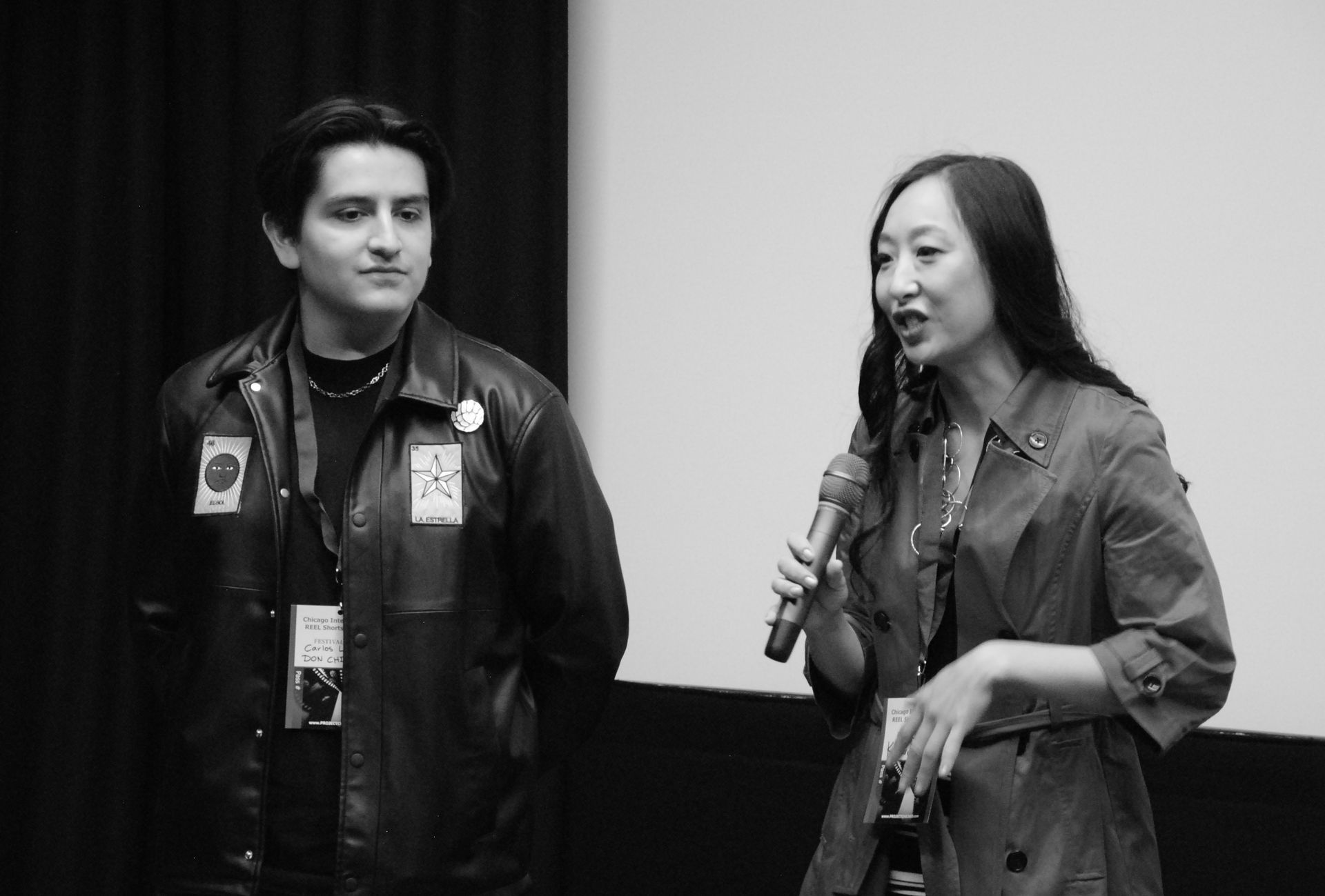 Two people on stage: a man in a leather jacket and a woman with a microphone, speaking in front of a screen.