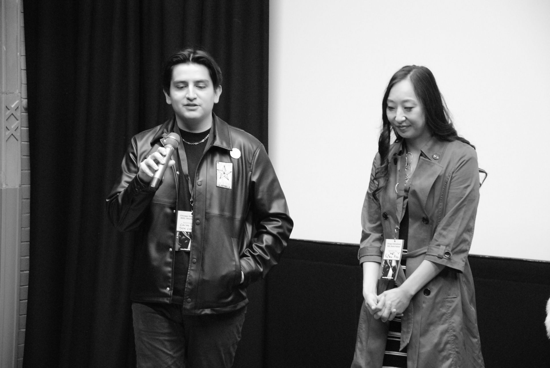 Man speaks into a microphone, woman smiles beside him. Both wear lanyards, standing in a room with a screen.