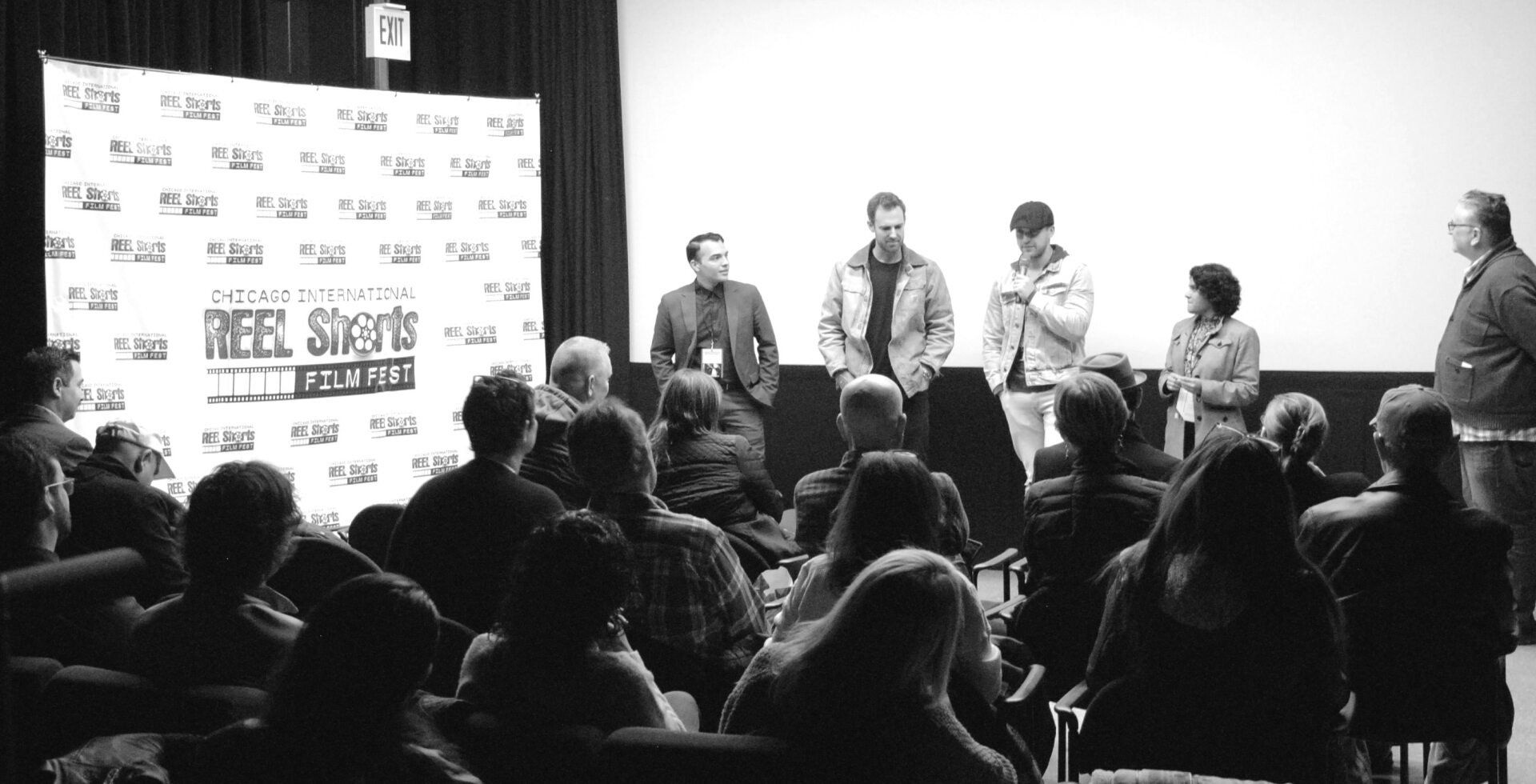 Panel discussion at film festival; speakers on stage, audience in foreground, black and white.