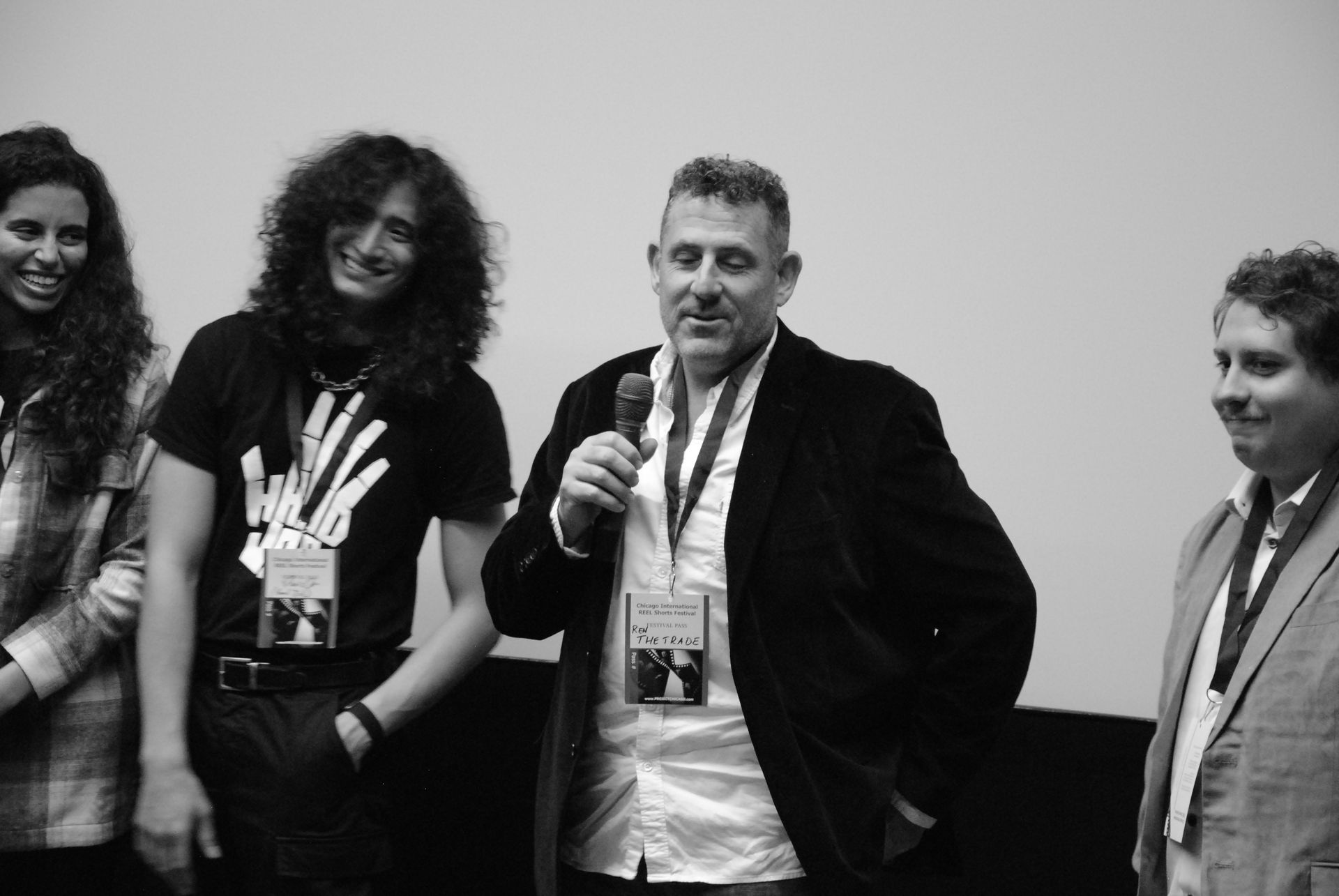 Group of four people at an event. Man with a microphone speaking. Others smiling. Black and white.