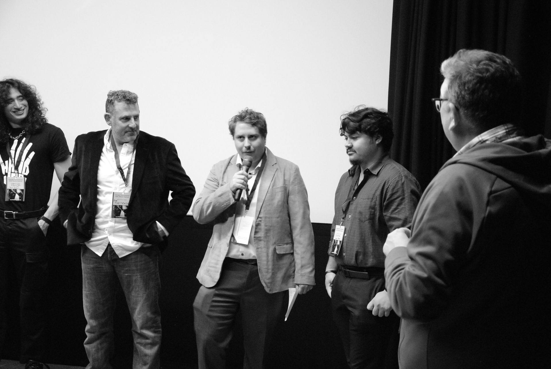 A group of five people standing on a stage, speaking to the audience. Black and white.