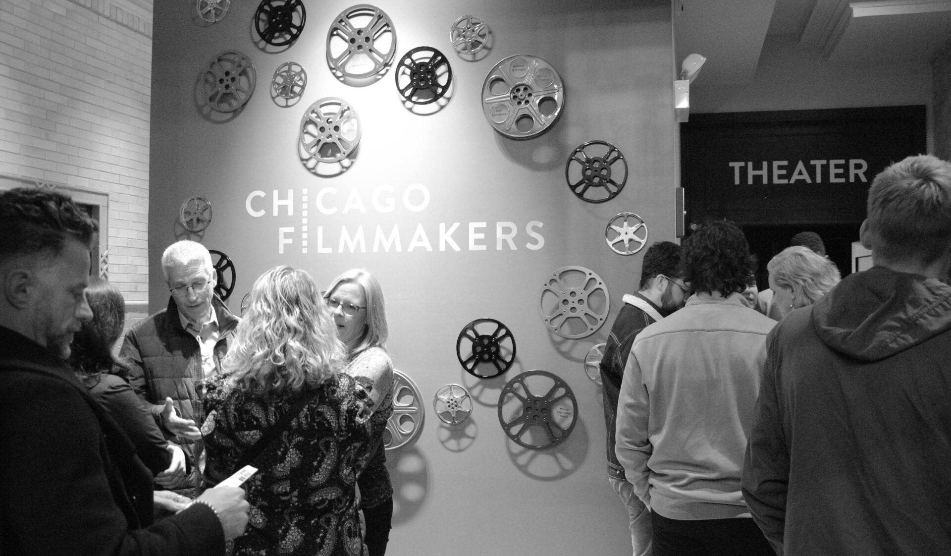 Black and white photo: People at Chicago Filmmakers event, near a wall with film reel art, theater entrance visible.