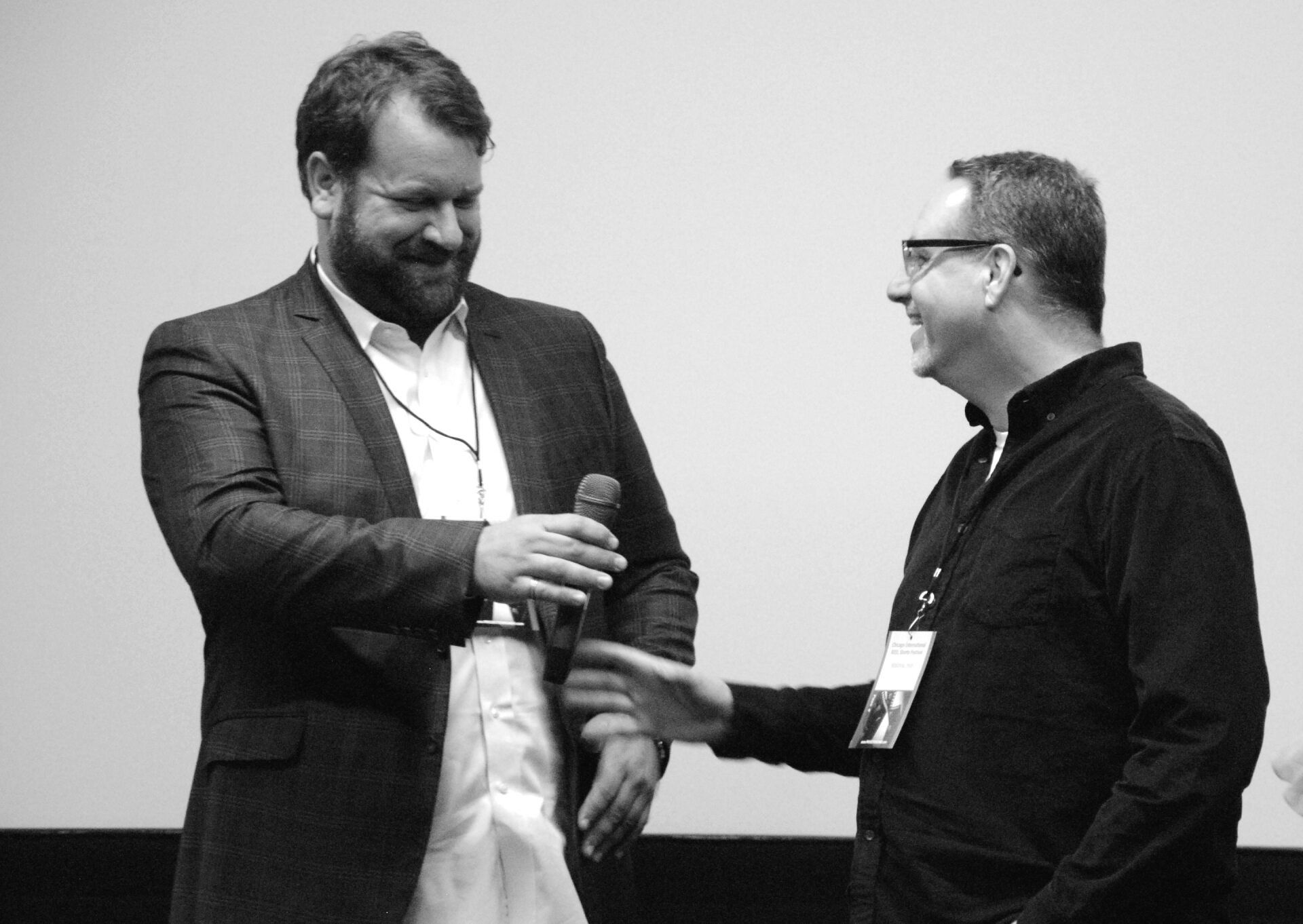 Two men shaking hands on a stage, one holding a microphone. Black and white.