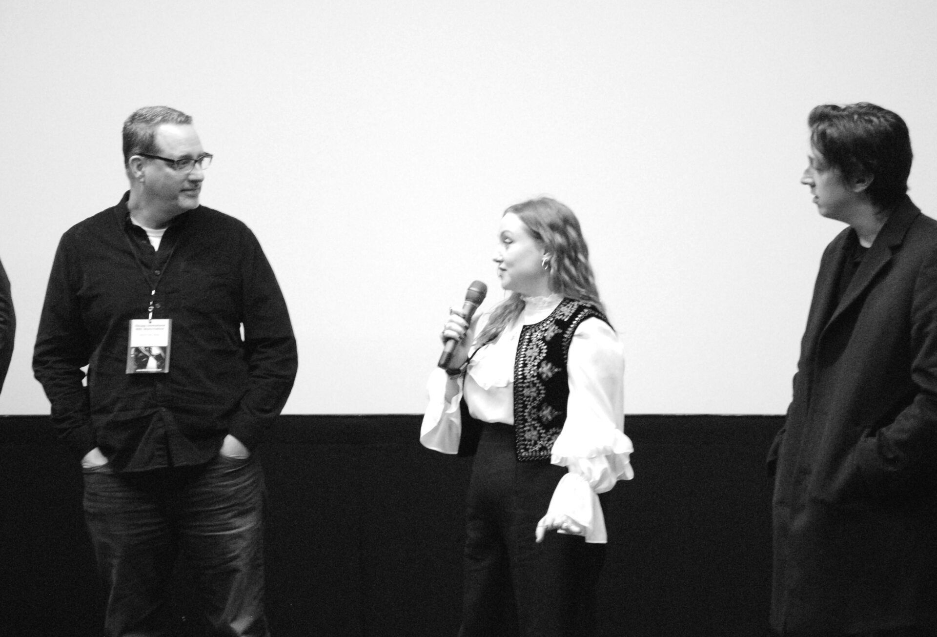 Three people on a stage, one with a microphone, speaking. Plain white background. Black and white photo.