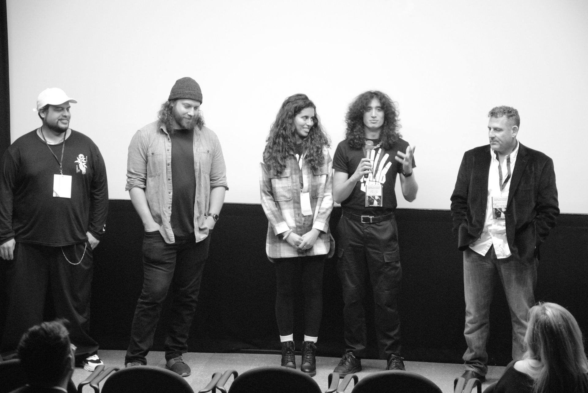 Panel of five people on a stage in front of a screen. They are speaking to an audience.