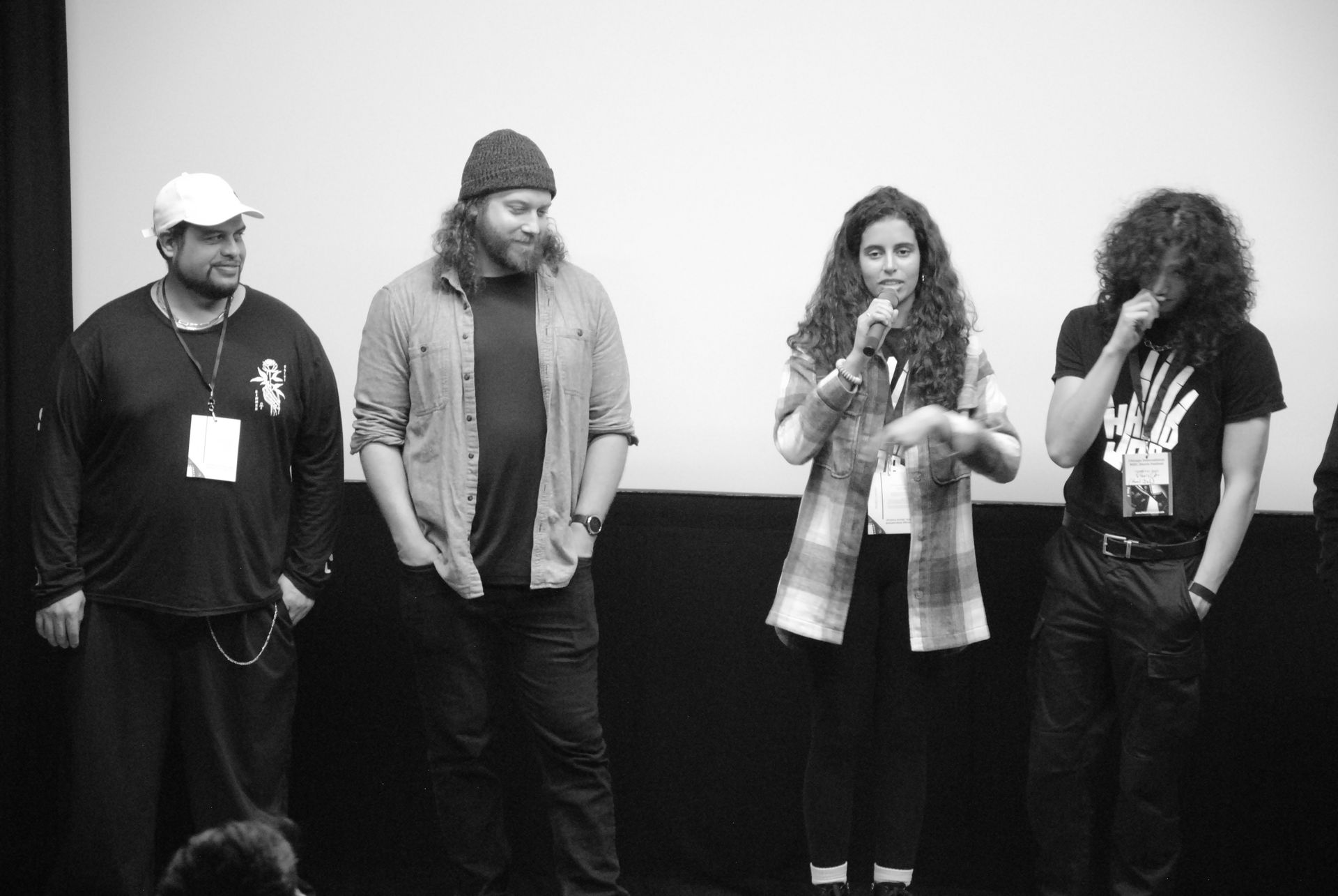 Four people on stage, one speaking with microphone, others listening; neutral background.
