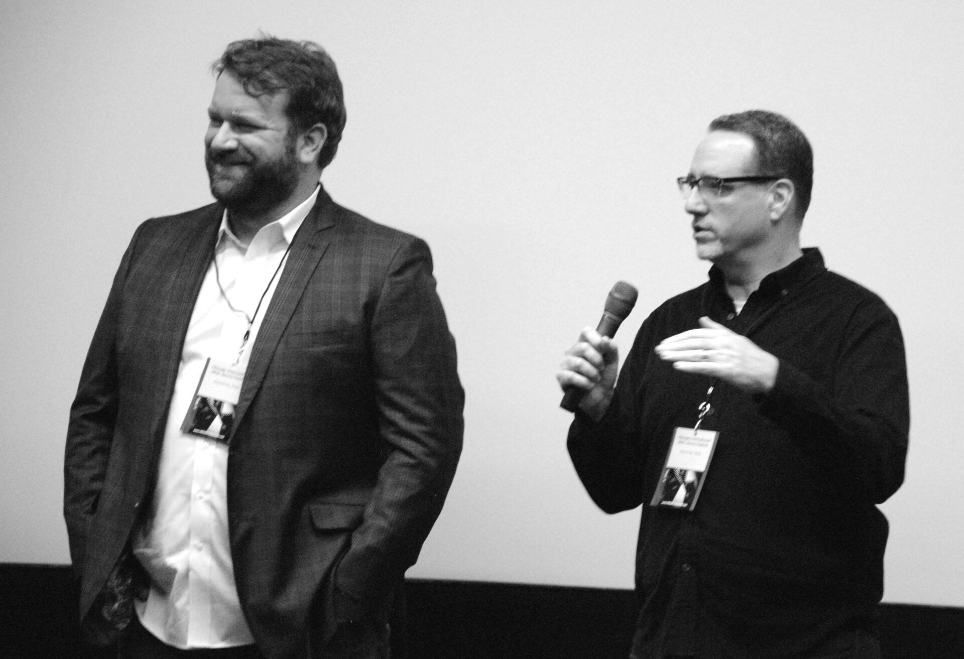 Two men on stage, one holding a microphone, the other smiling, both wearing lanyards. Black and white photo.