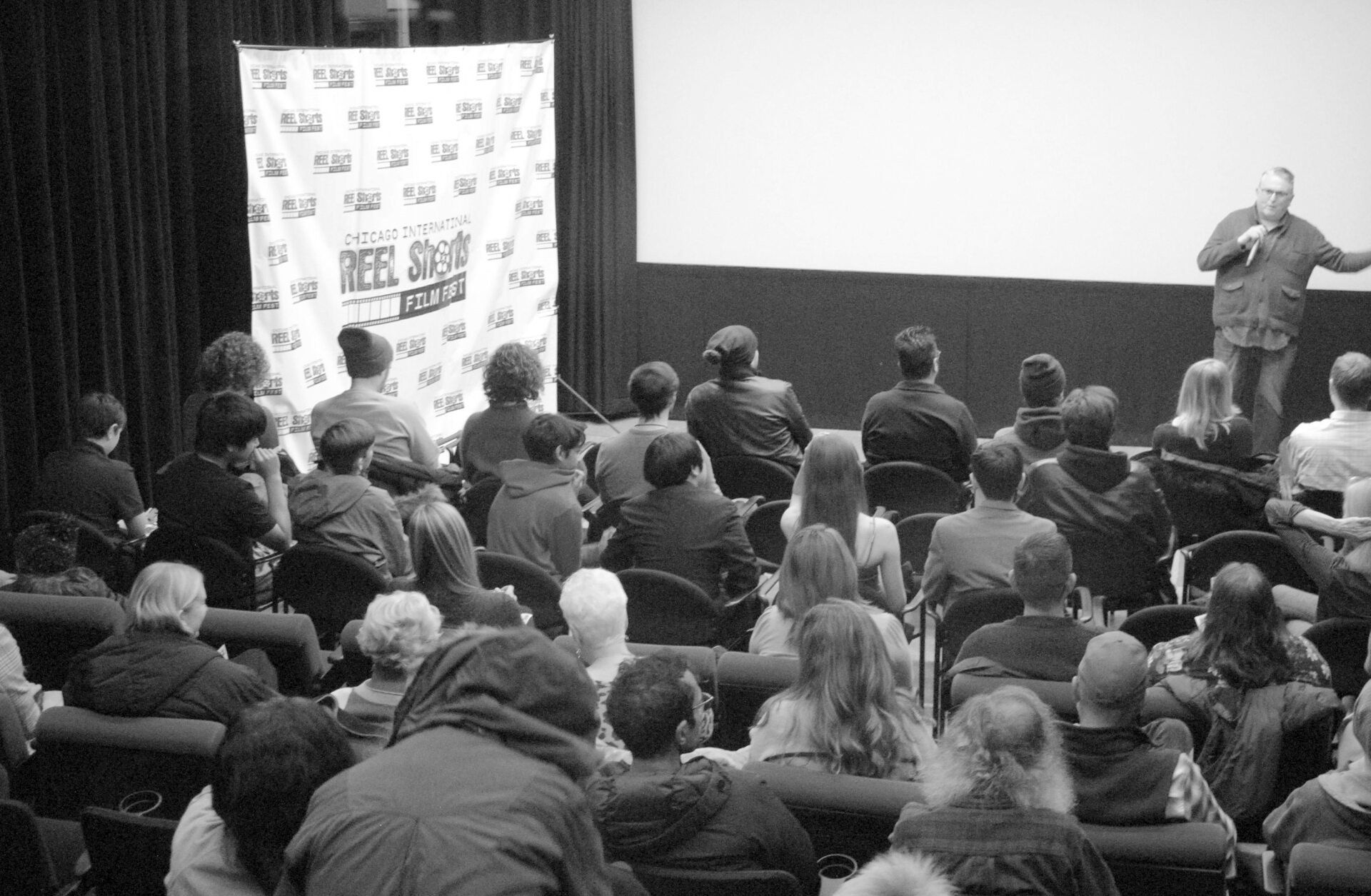 Audience watching a man speaking on stage, screen behind him. Event at a theater. Black and white.