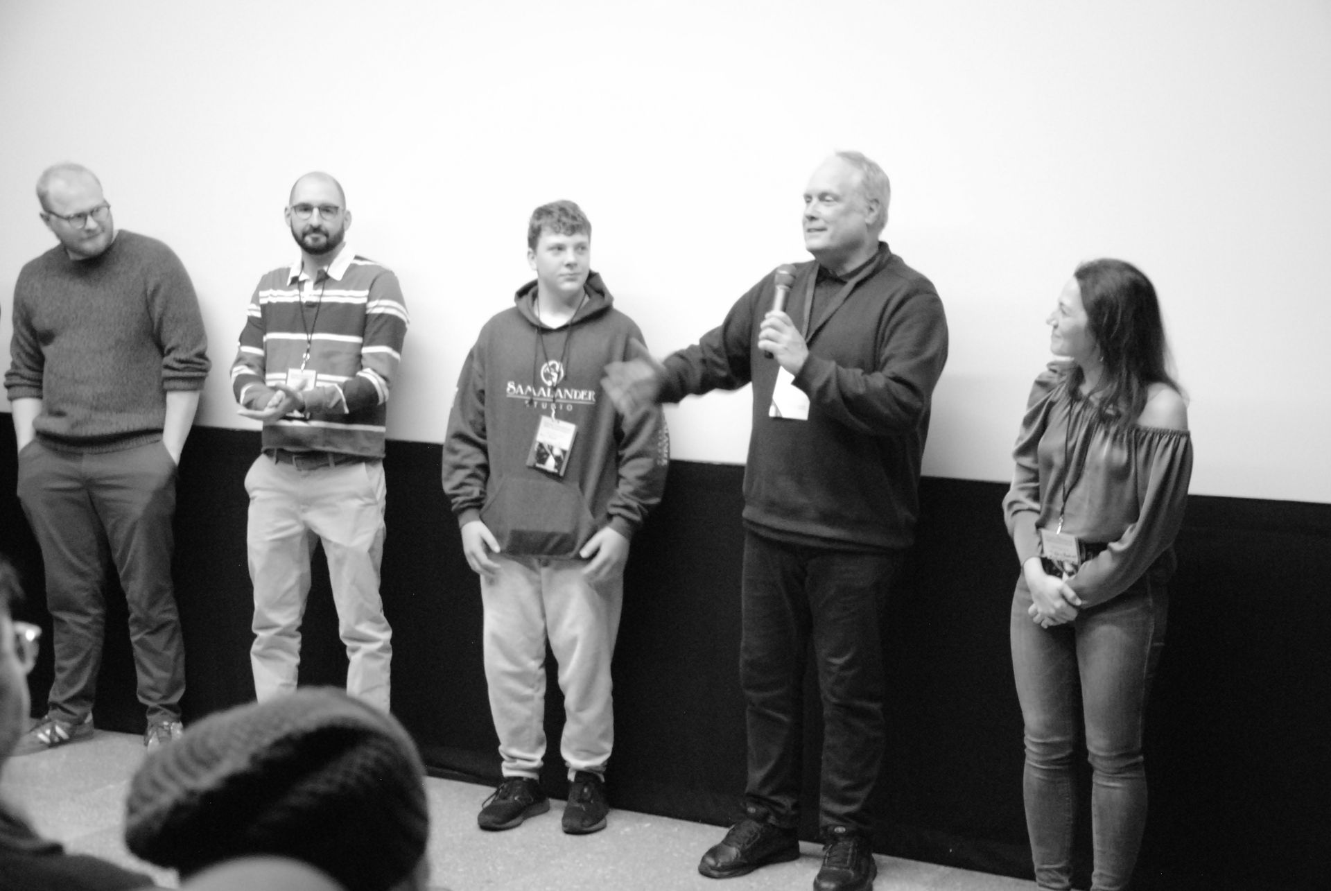 Group of six people at a conference. A man with a microphone speaks, gesturing toward a young man in a hoodie.