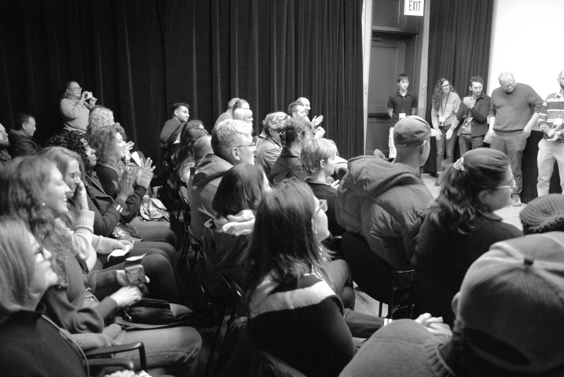 Audience applauds performers on stage in a dimly lit room; black backdrop; many people seated.