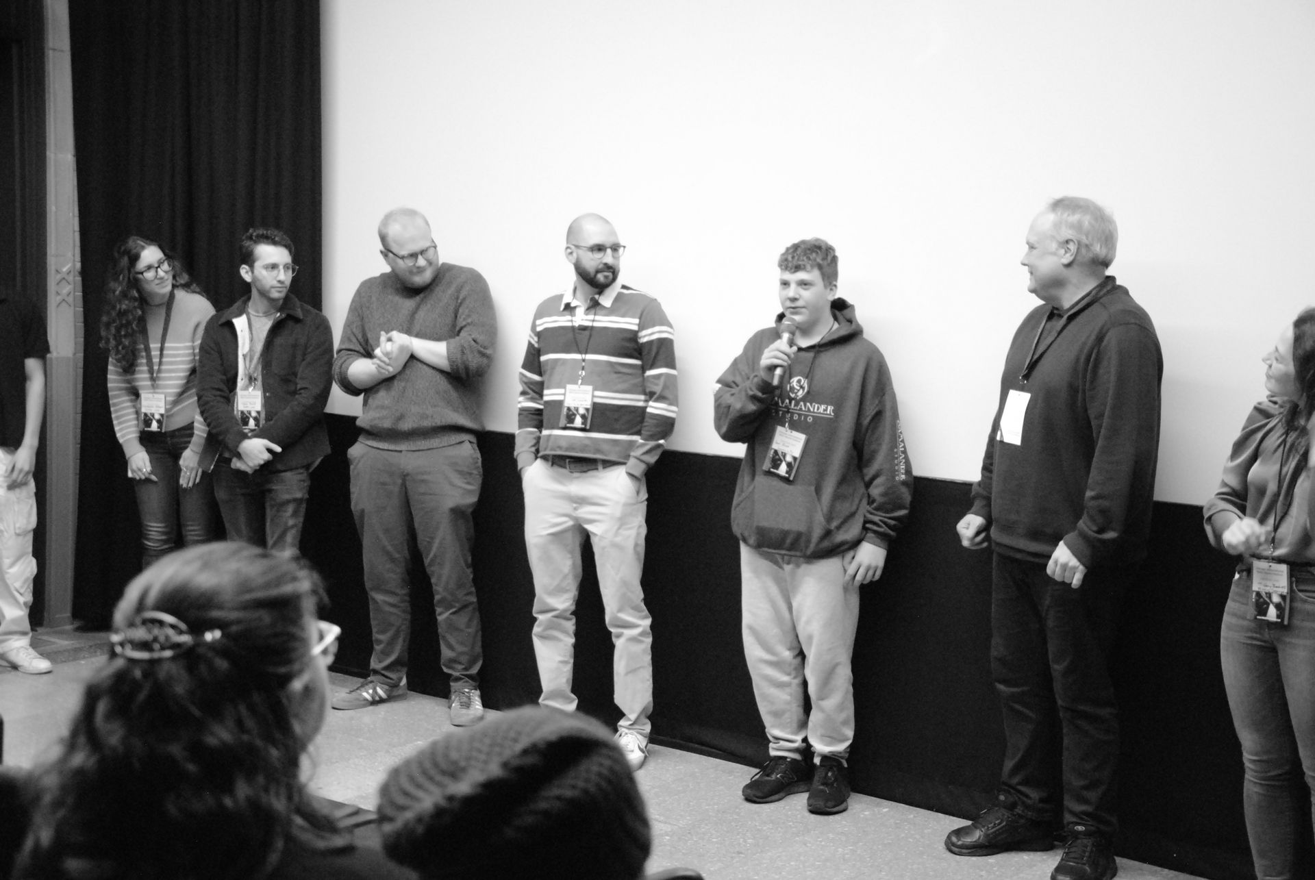 Group of people standing in front of a screen. One person speaks into a microphone. Black and white.
