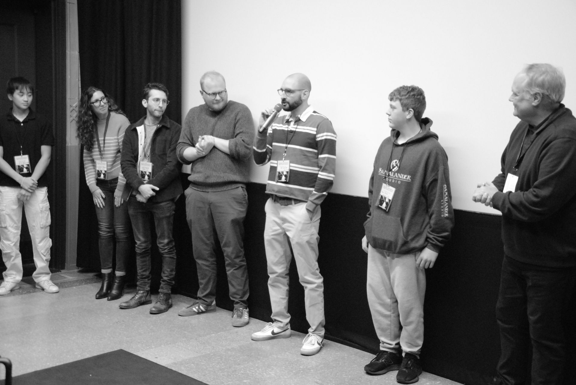 Group of people standing on a stage, some with microphones, in front of a screen, appearing to be speaking.