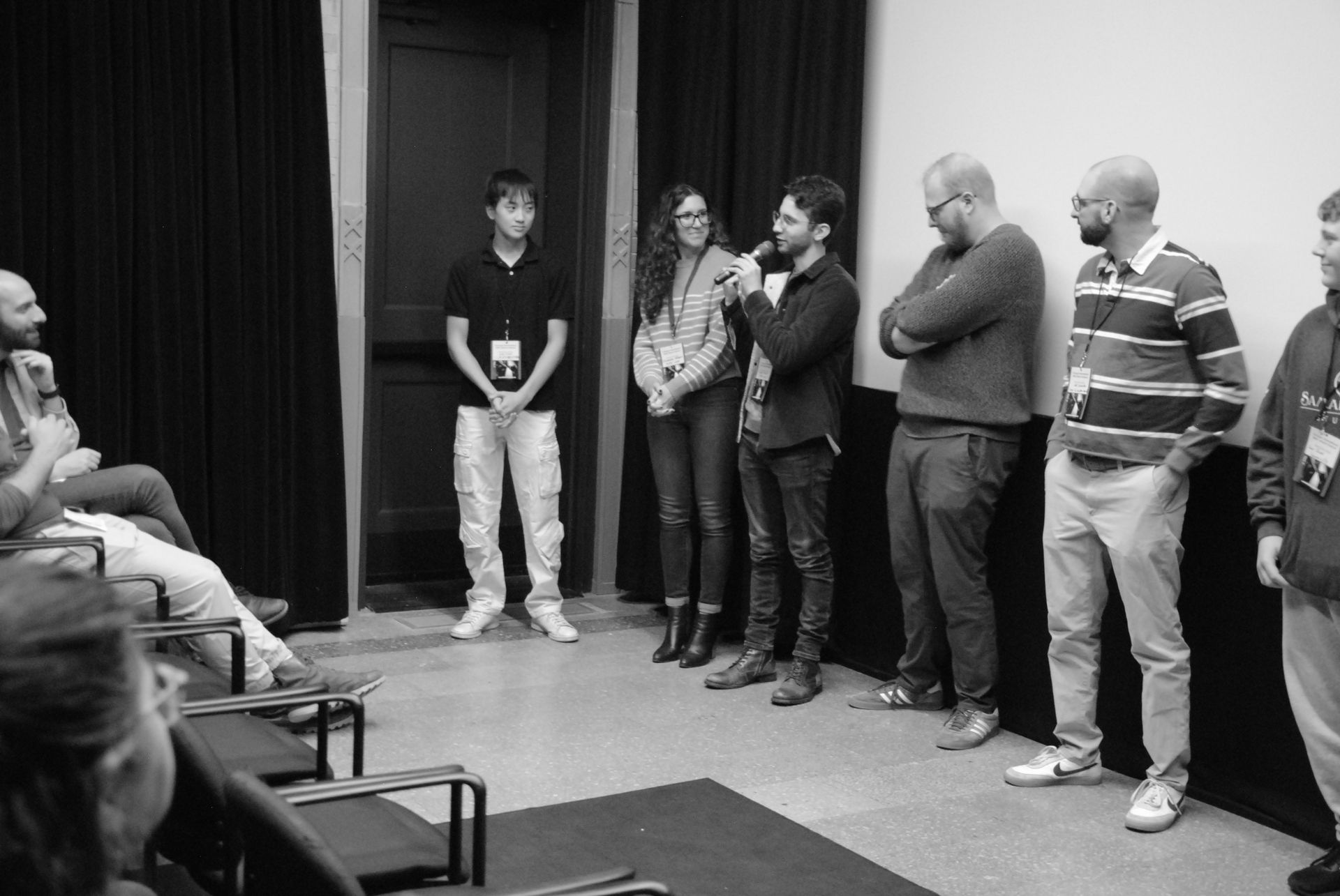 People standing and sitting in a room, possibly at a panel or discussion. One person speaks into a microphone.