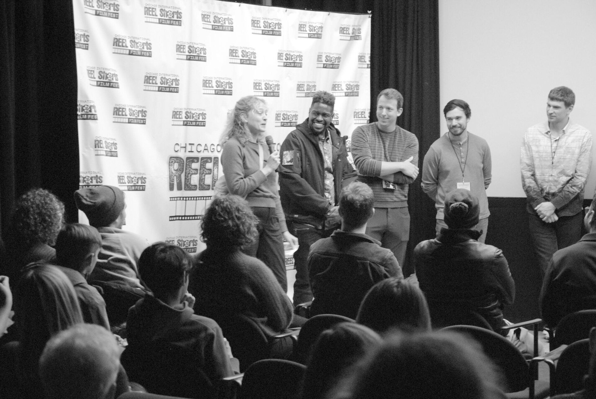 Panel of five speakers on a stage at a film festival, addressing an audience. Black and white.