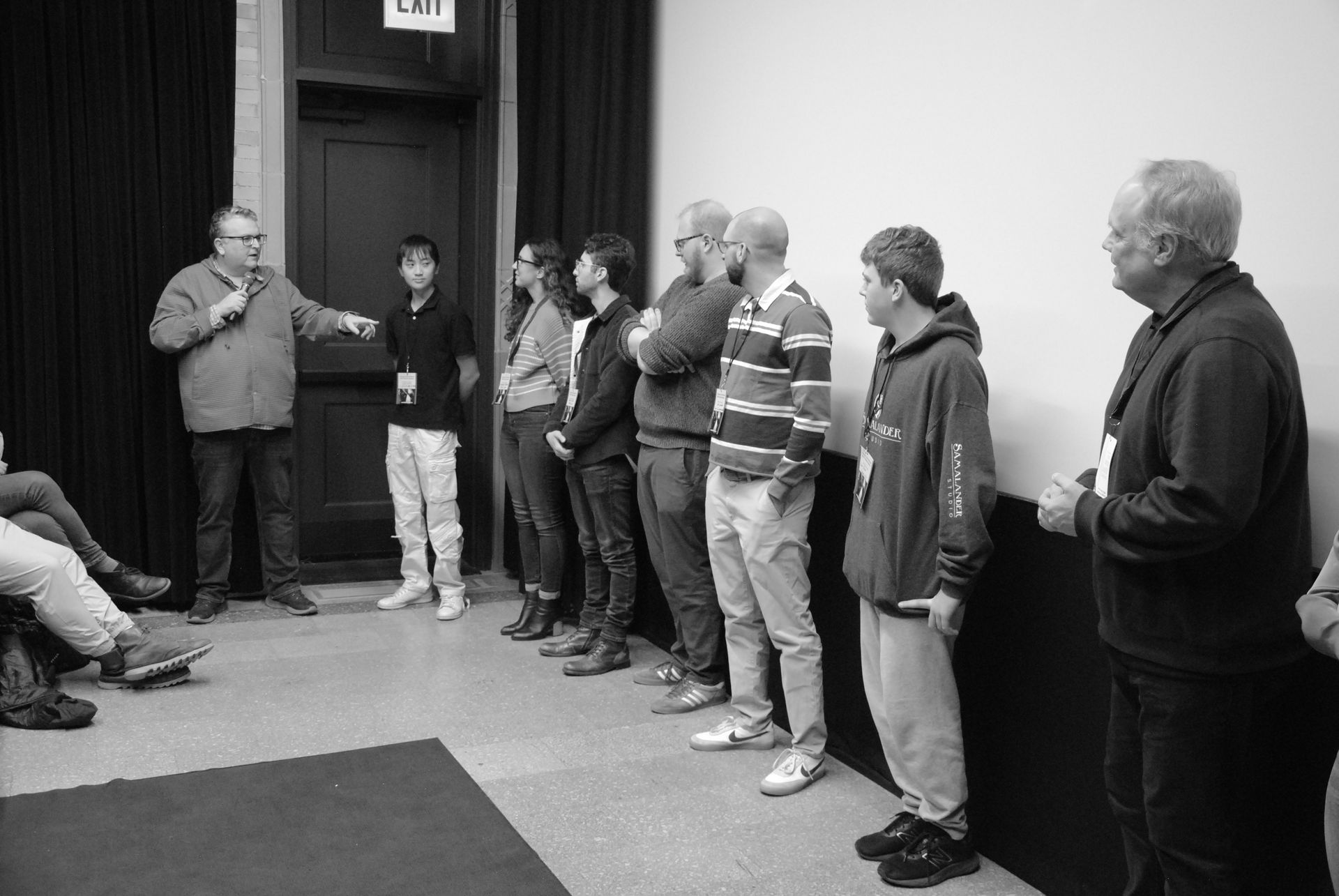A group of people in a room; a man is speaking and gesturing while others listen, standing or sitting.