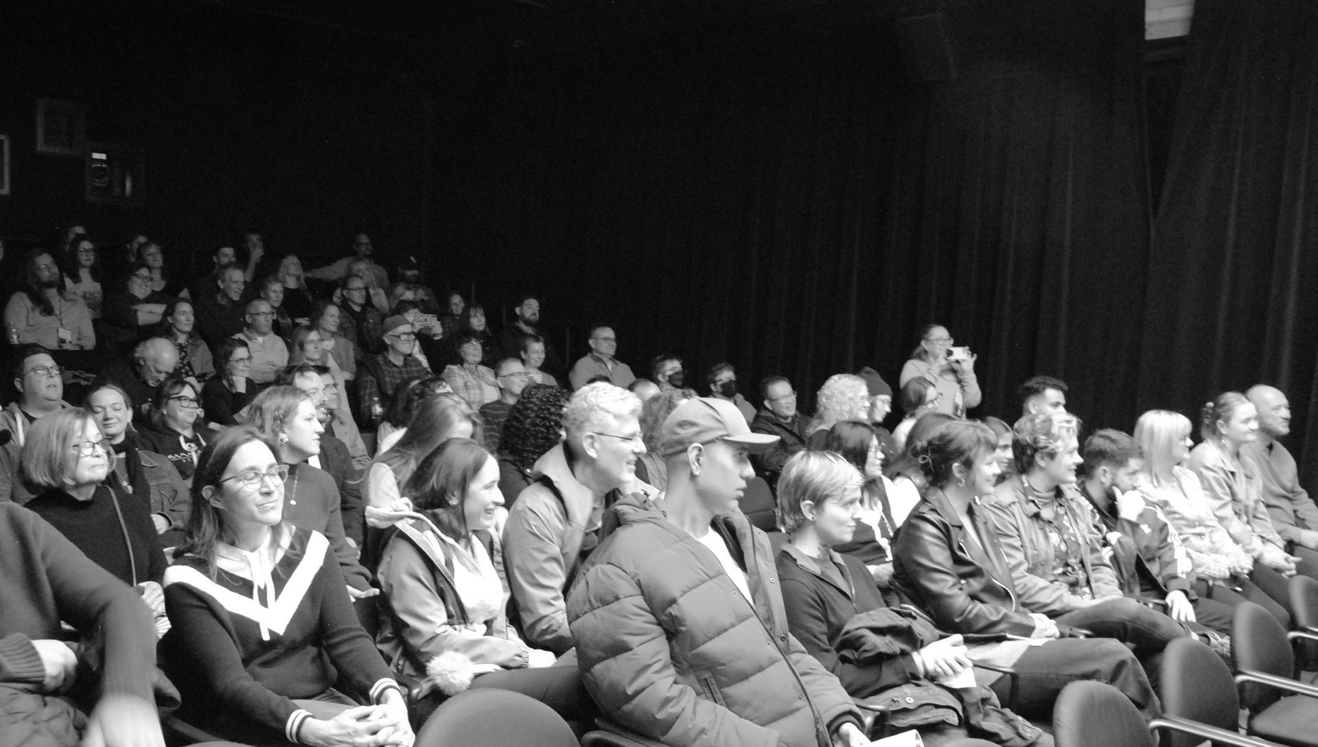 Audience seated in a darkened theater watching a performance. Many people are visible.