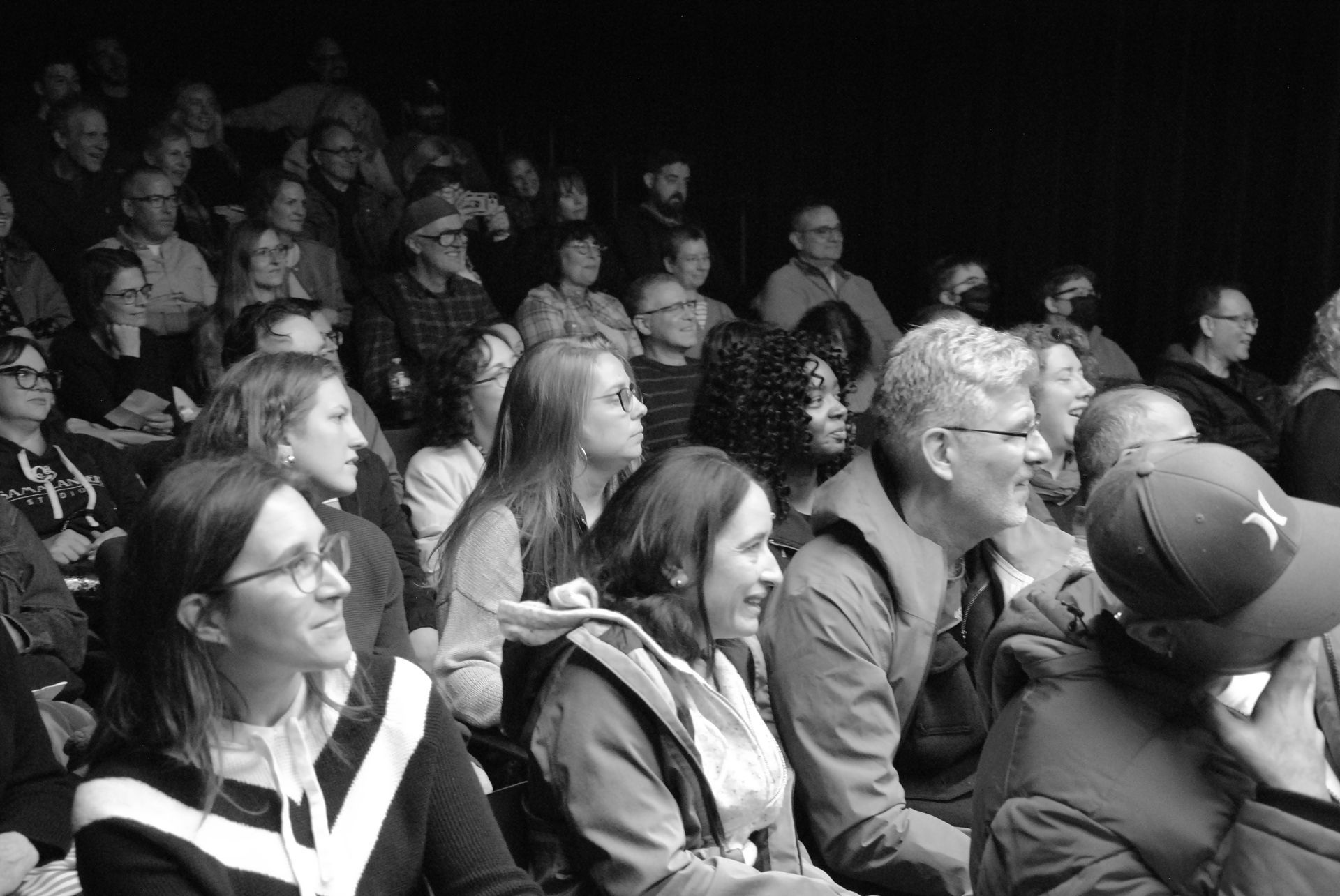 Audience in a theater; people watching, some smiling. Dark setting.