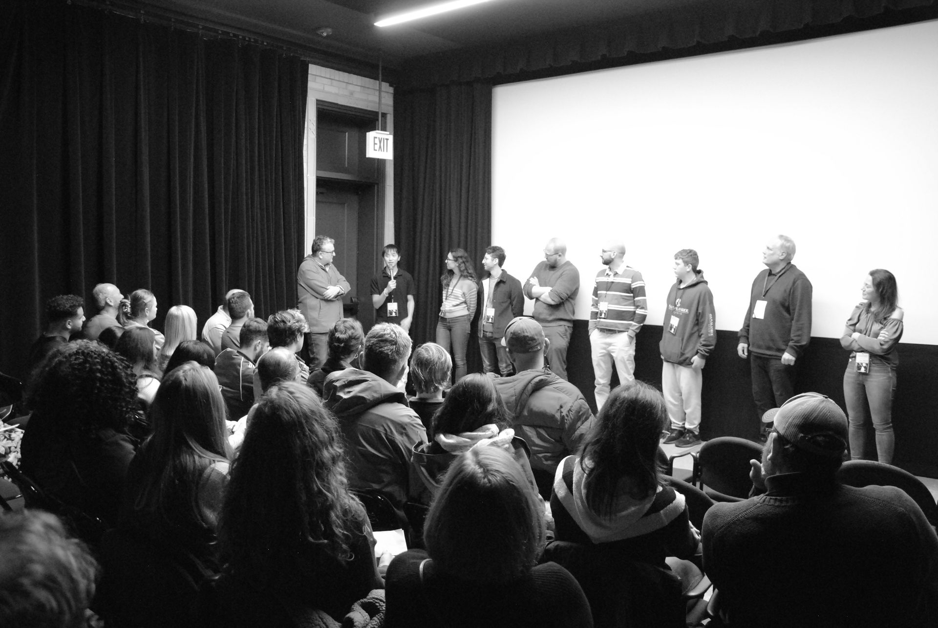 A panel of people stands on a stage facing an audience in a theater setting.