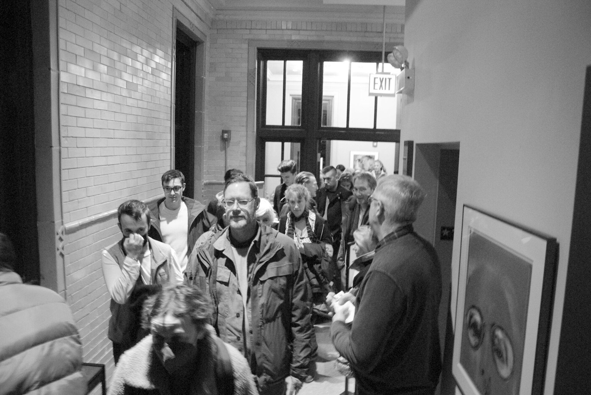 People walking down a hallway with white brick walls, toward a door. Mostly men, some looking forward, others towards each other.