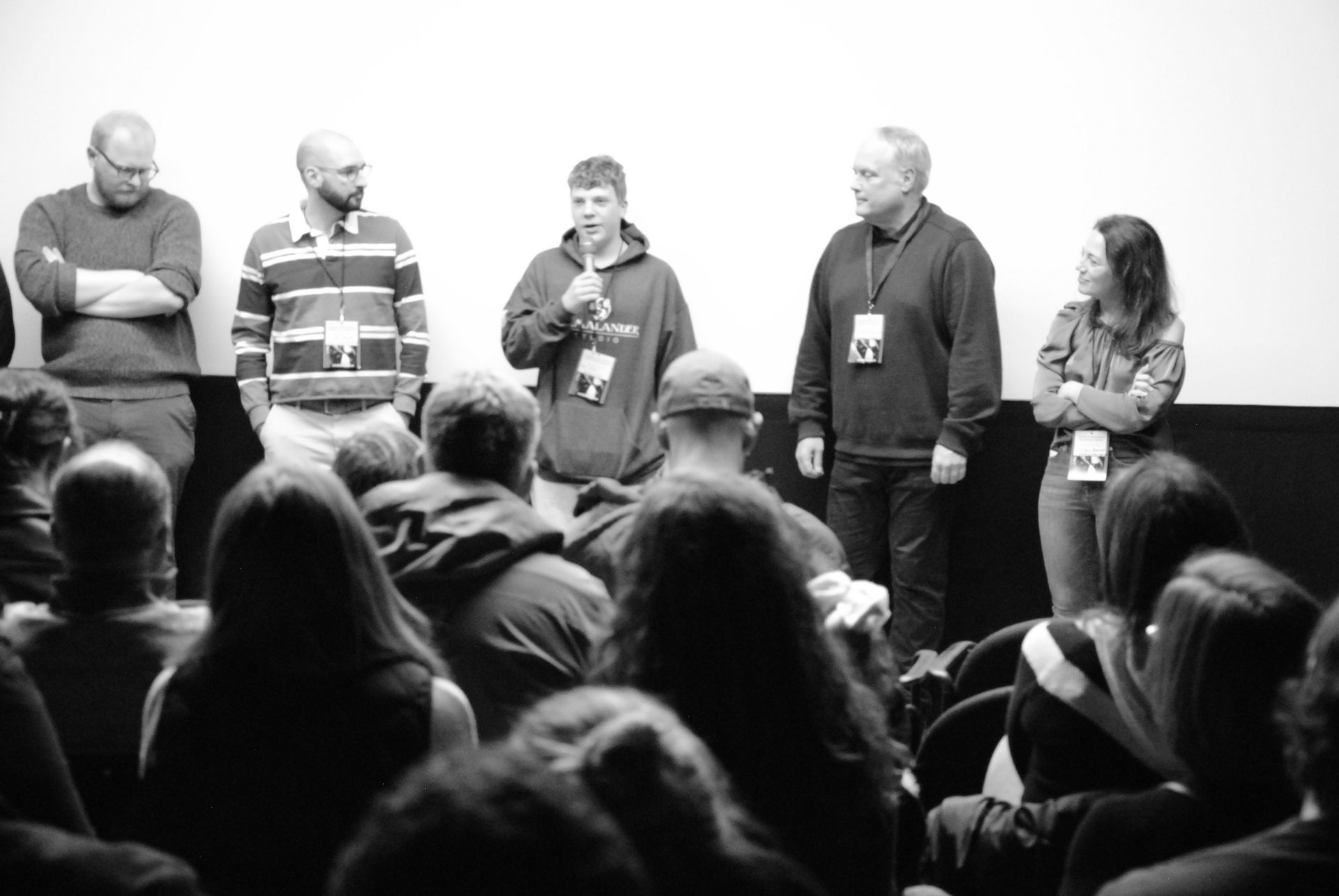 Panel of five people addressing an audience in a dark room. One holds a microphone, others stand.