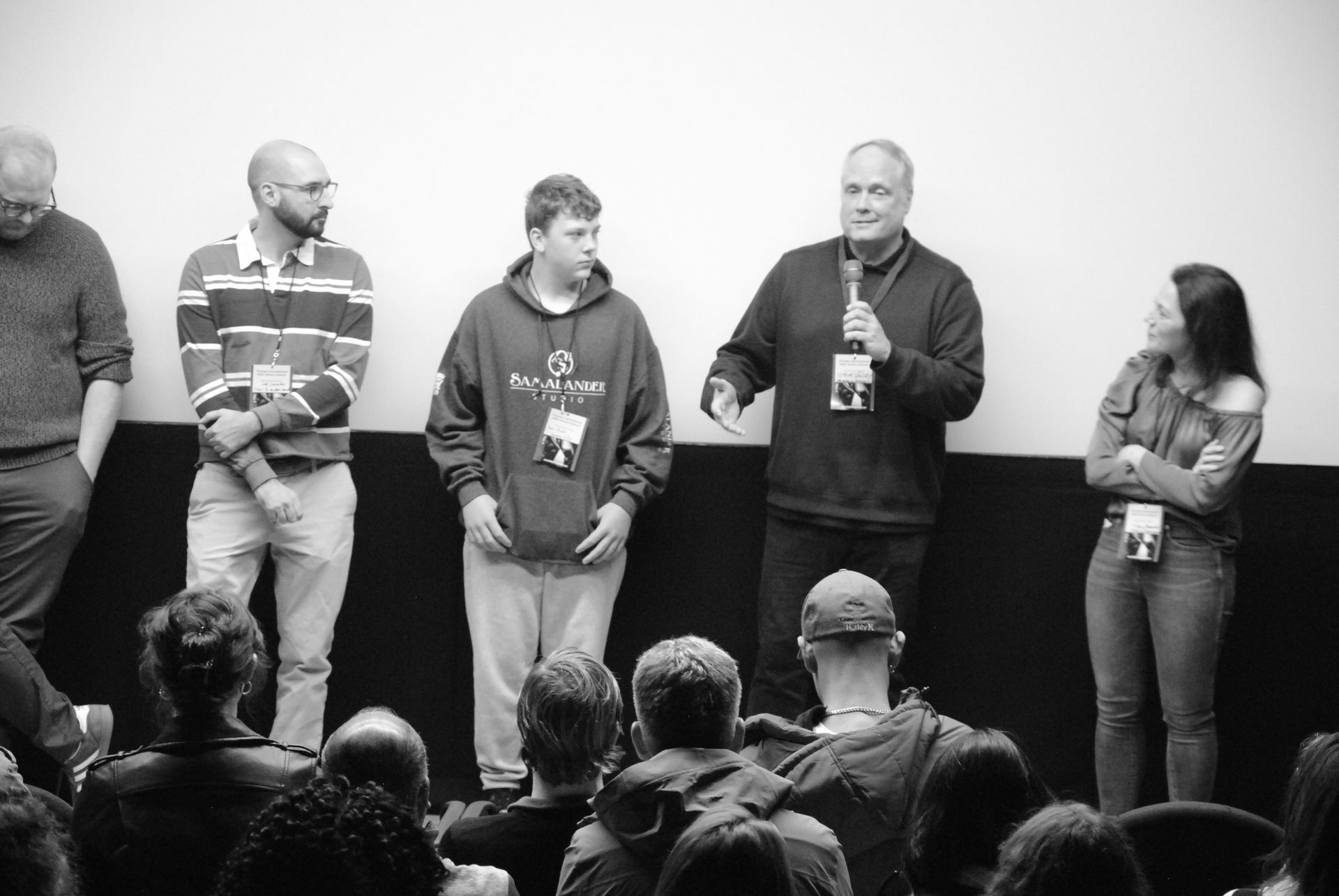 Panel of speakers at an event, standing in front of an audience. Man with a mic speaks, other people listen.