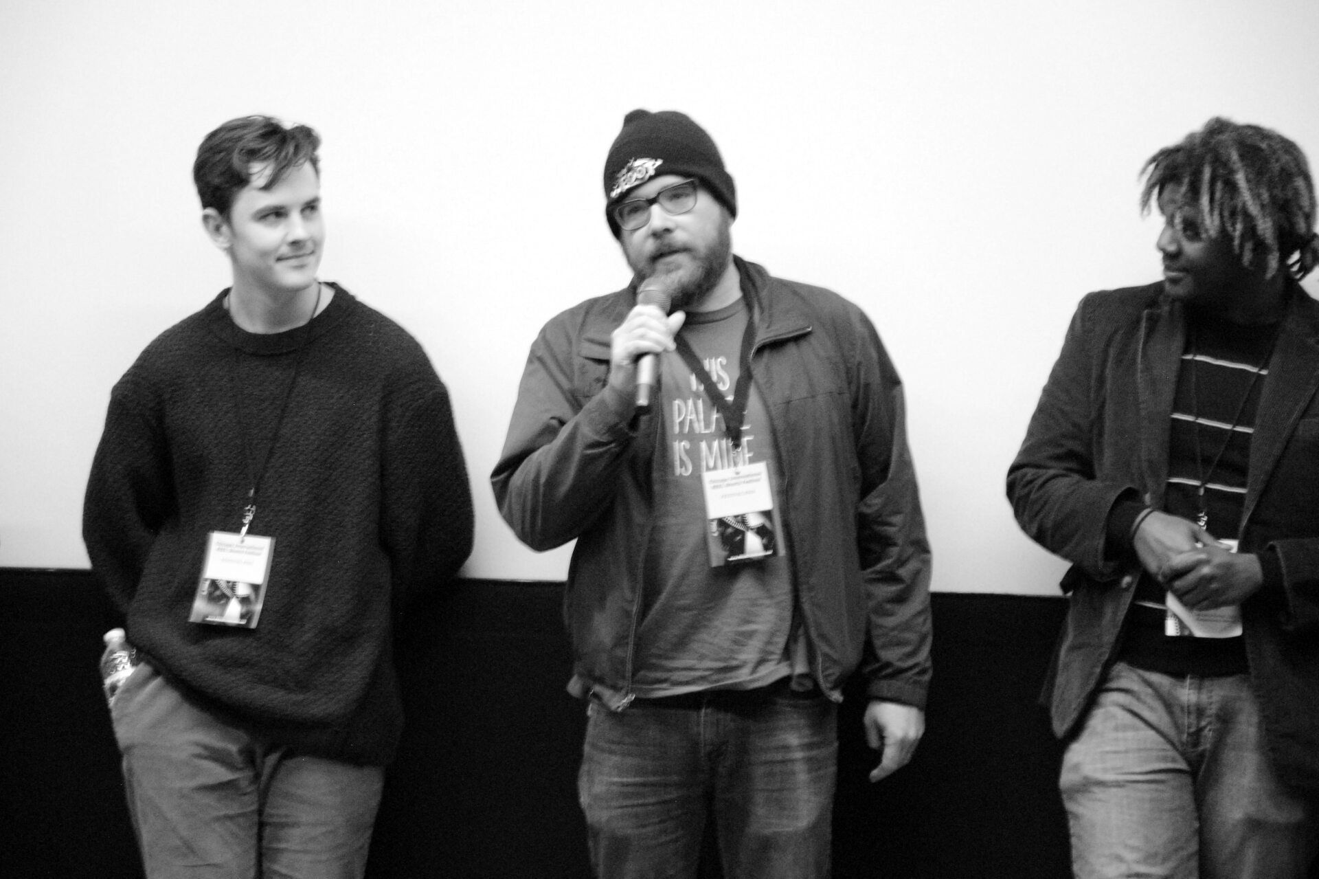 Three people on stage at an event, one with a microphone speaking, all wearing badges. Black and white.