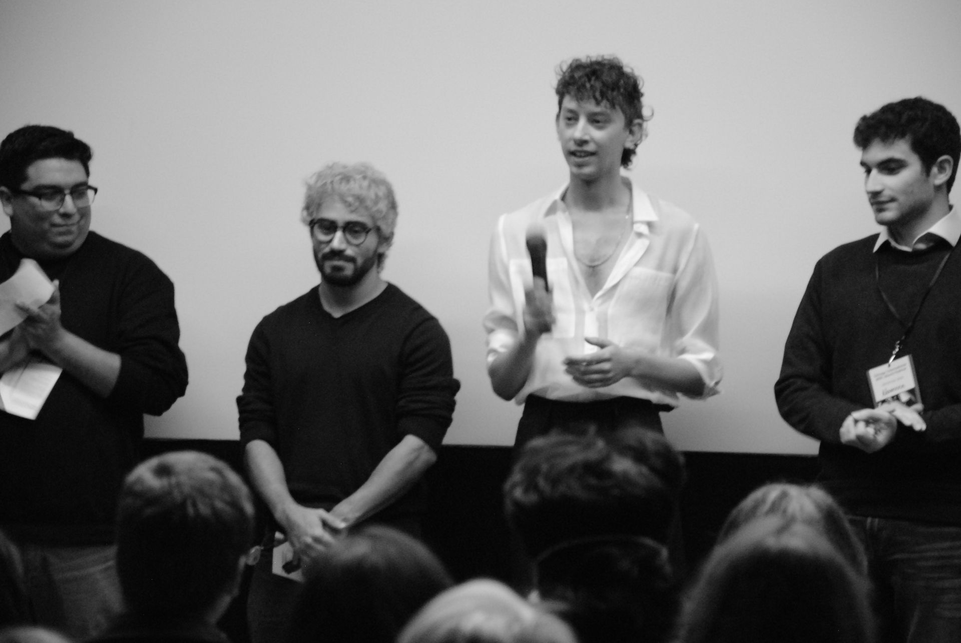 Four people on stage, one speaking into a microphone, facing an audience. Black and white photo.