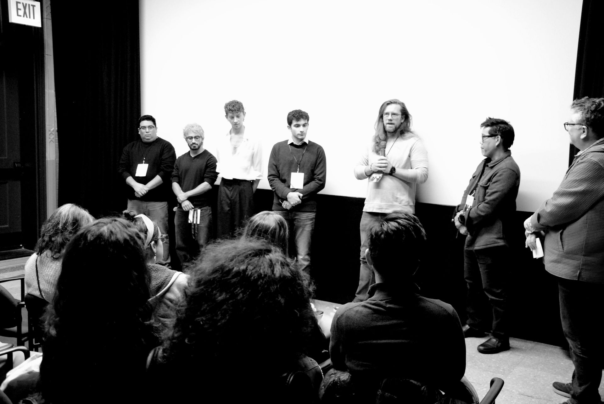 A panel stands before an audience in a theater. Speakers hold microphones, one has long hair, and all wear name tags.