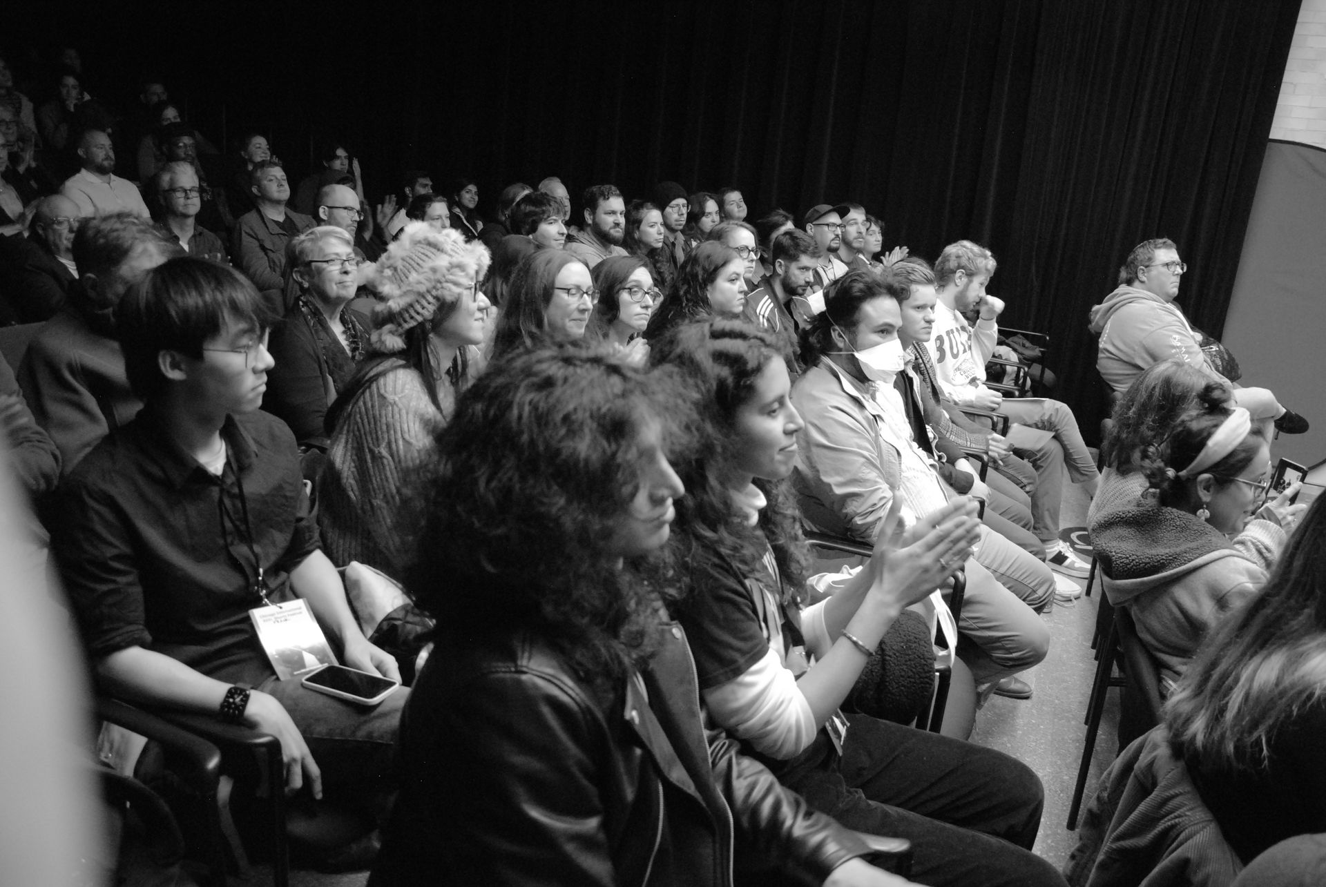 Audience in a theater. People of diverse ages and appearances sit, some clapping. Black and white photo.