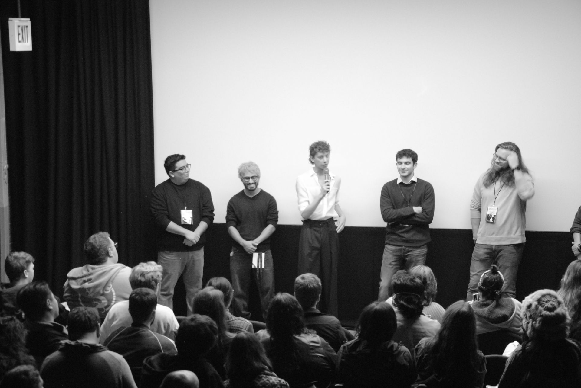 Panel of five men on a stage facing an audience. Men stand in front of a white screen. Black and white photo.