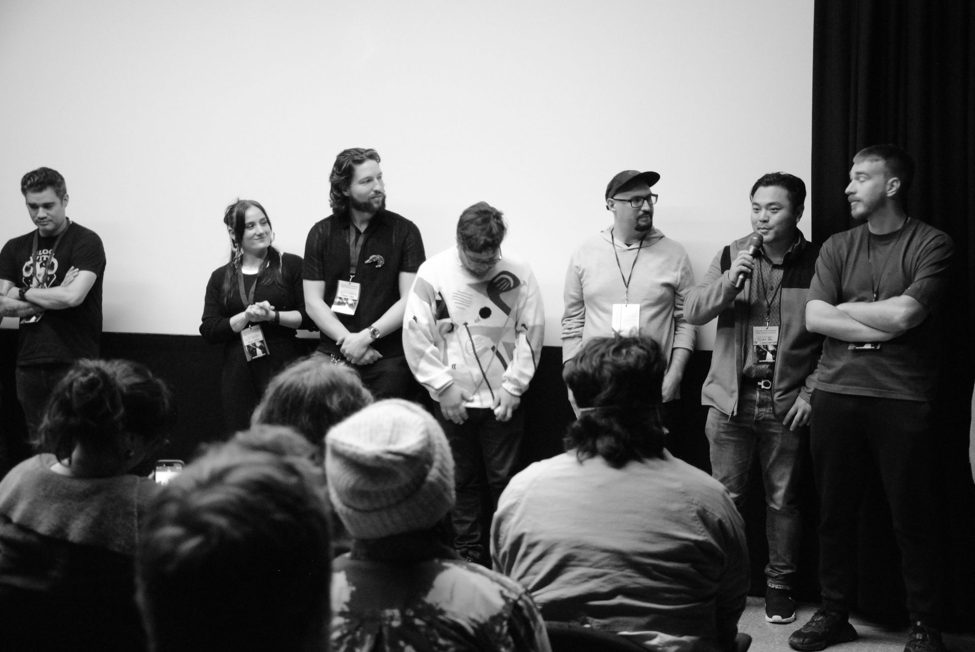 Panel of people on a stage in front of an audience. Several are holding microphones, wearing lanyards, and smiling.