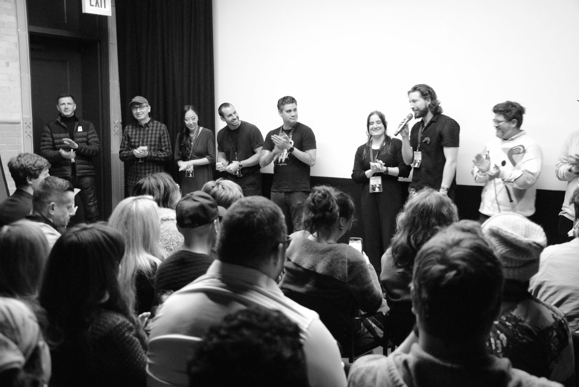 Film crew onstage applauds after screening; audience in foreground; theater setting.