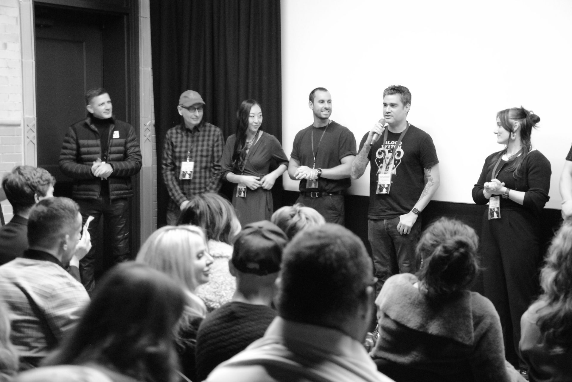 A group of people stand on a stage, addressing an audience. They appear to be panel speakers. Black and white.