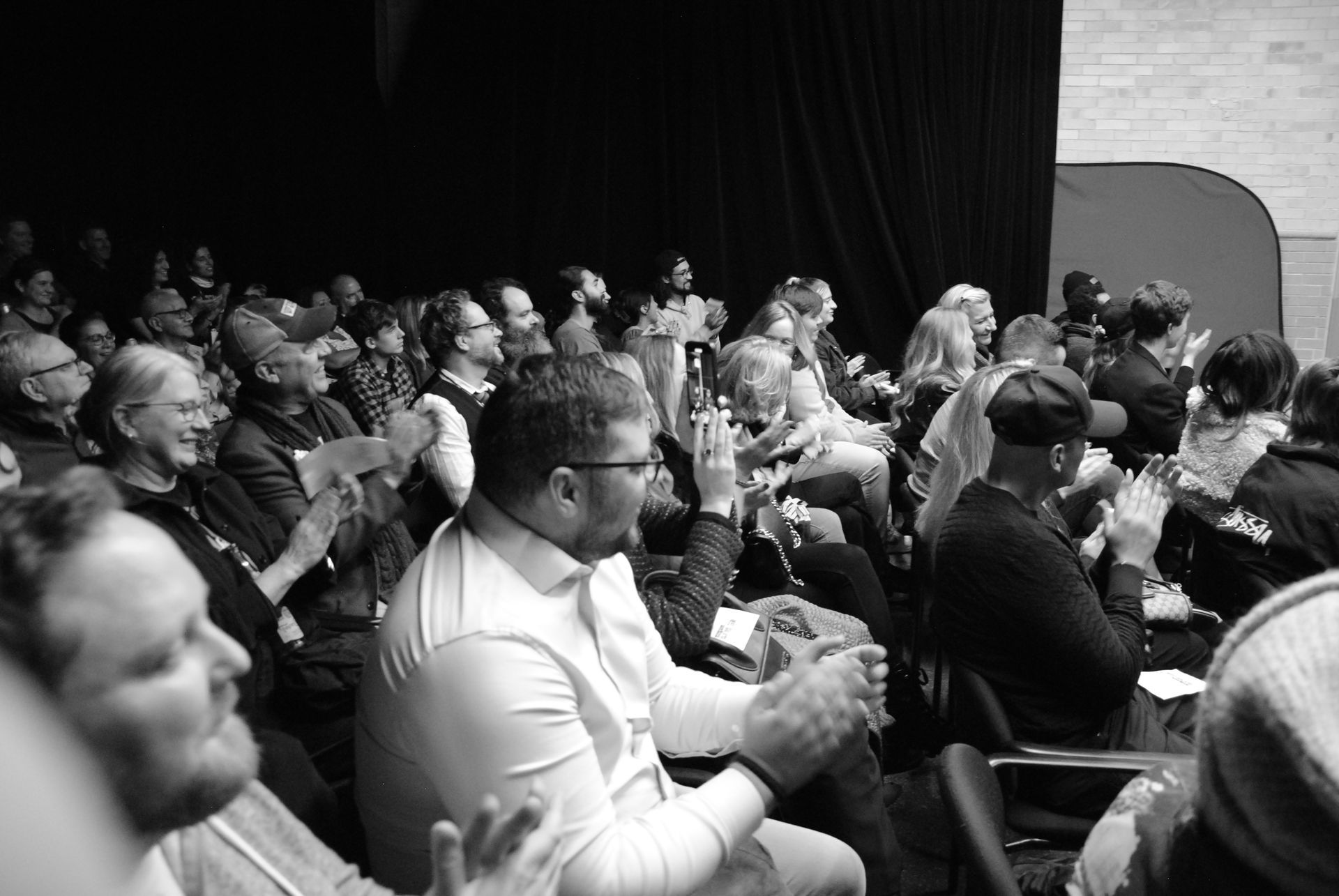 Audience applauds at an event. People of various ages and ethnicities seated in rows, facing forward.